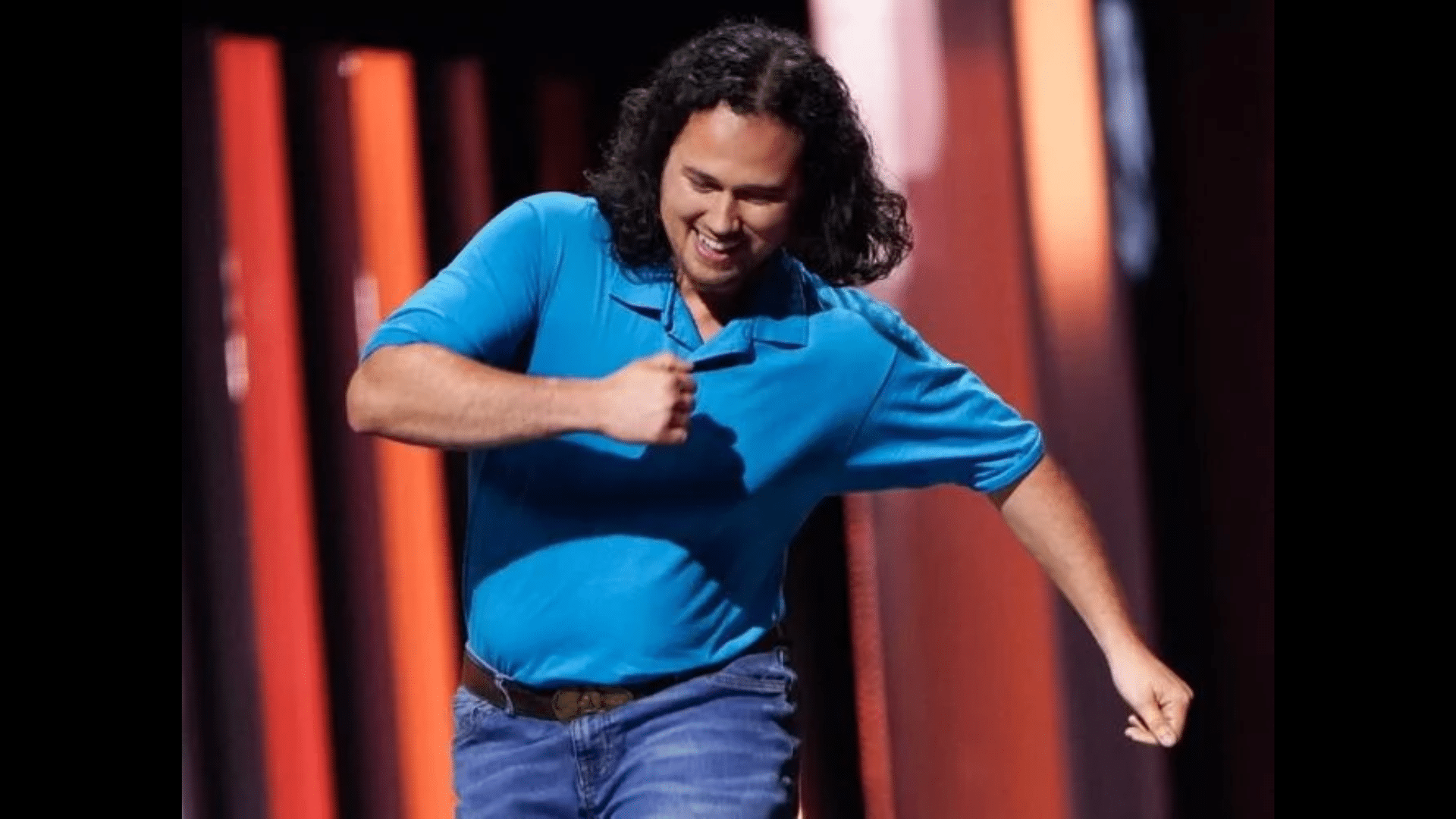 Zeb Ross with long curly hair in a blue polo shirt and jeans is smiling and dancing on a stage.