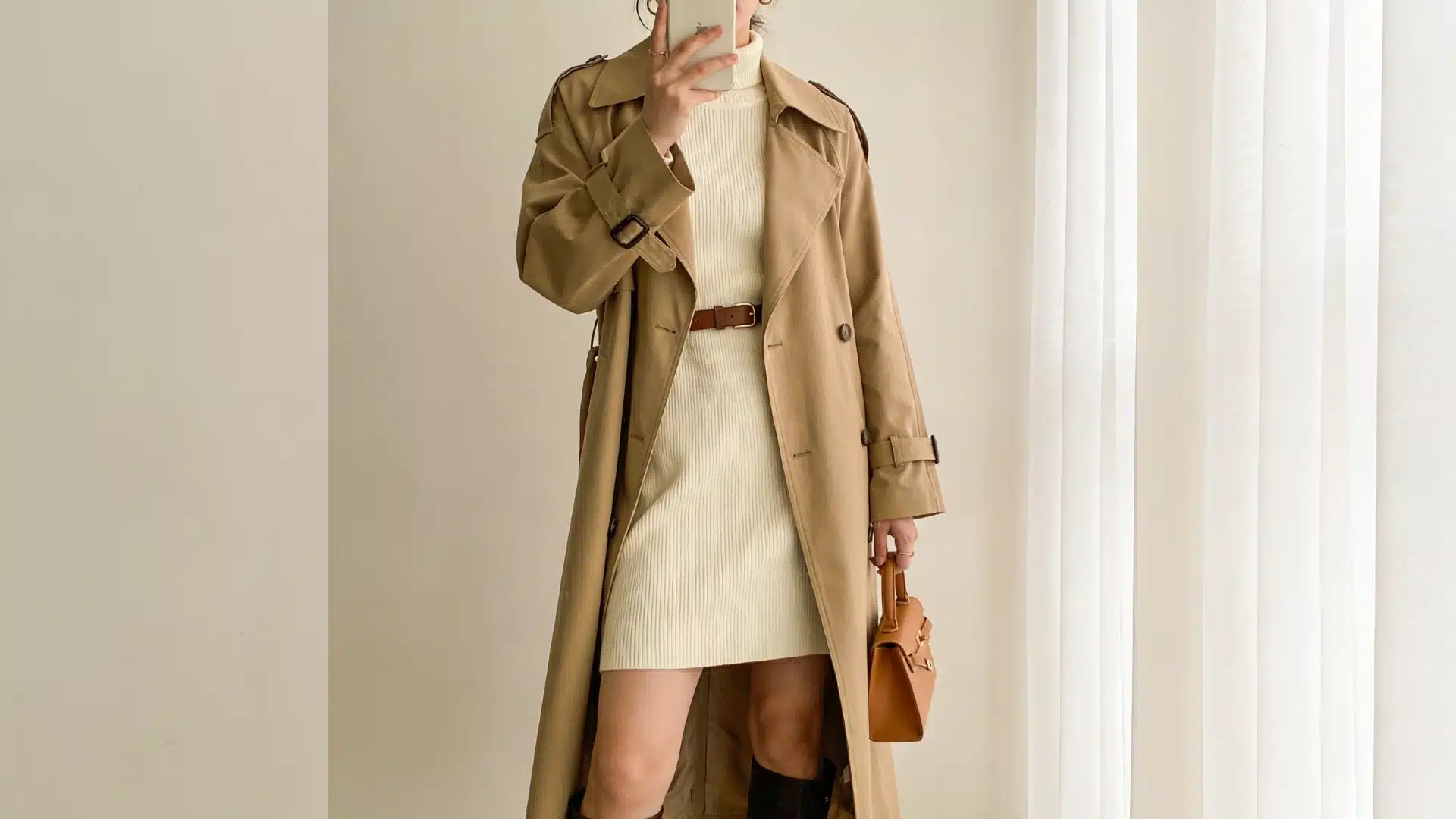 Woman in a beige trench coat, cream knit dress, brown boots, and a small leather handbag taking a mirror selfie with a smartphone.