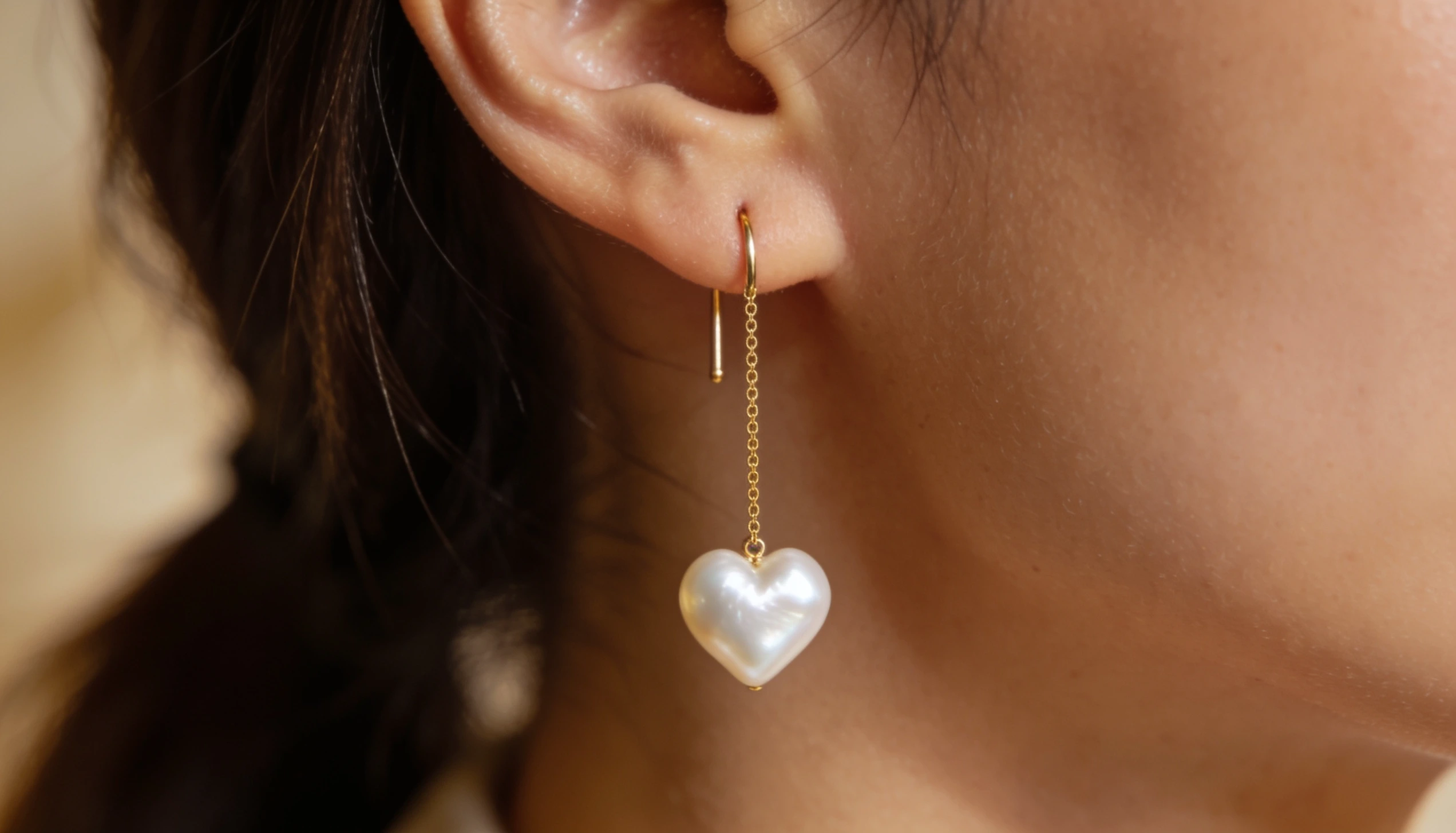 Close view of an ear wearing a gold threader earring with a delicate chain and heart shaped pearl pendant in warm natural light
