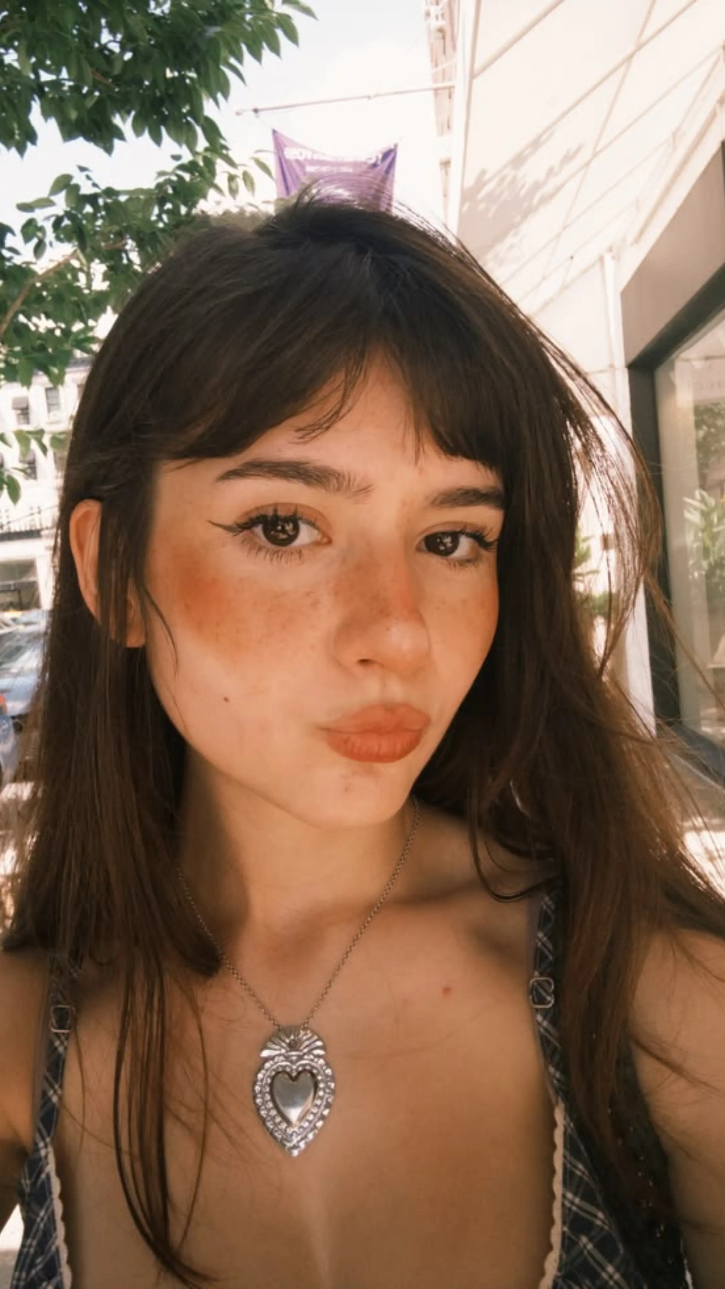 A young woman with dark hair and baby bangs wears a silver heart pendant necklace, looking forward on a sunny street.
