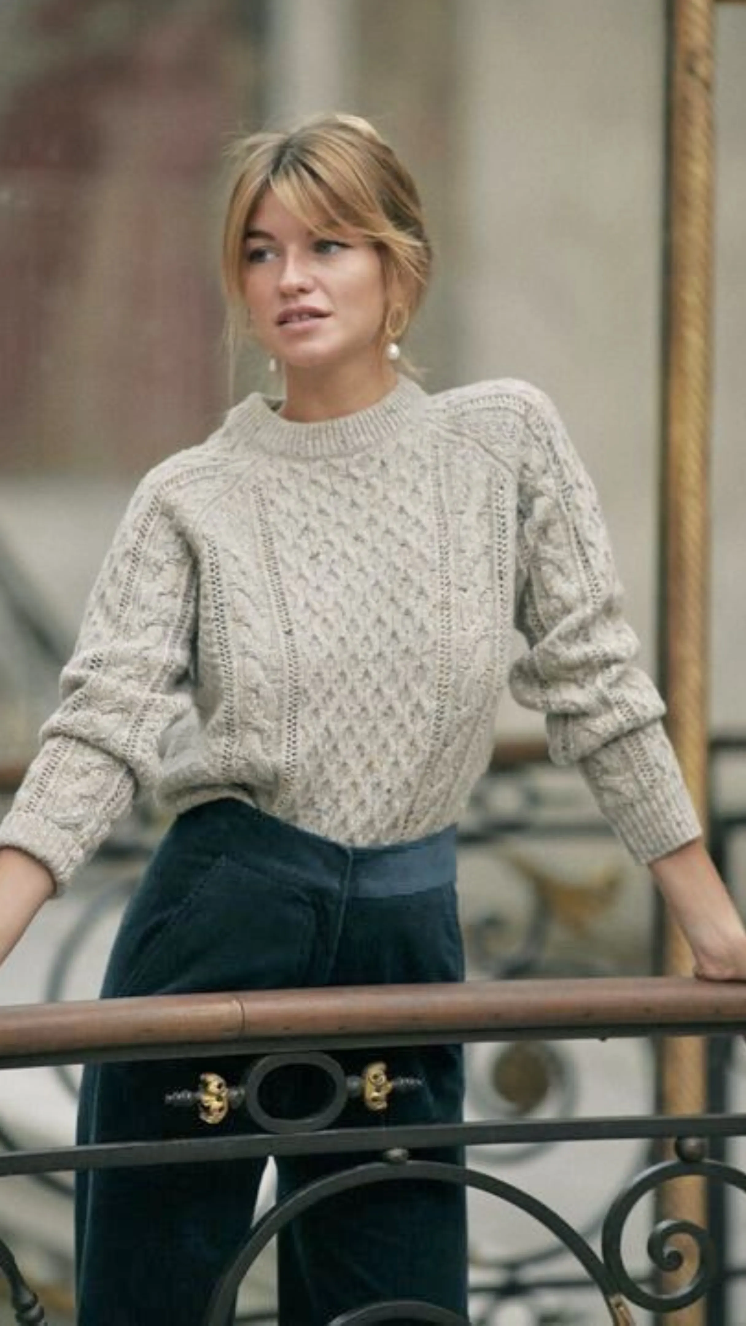 A young blonde woman with soft bardot bangs wearing a textured cable-knit cream sweater and dark trousers leans against a wrought iron balcony railing outdoors.