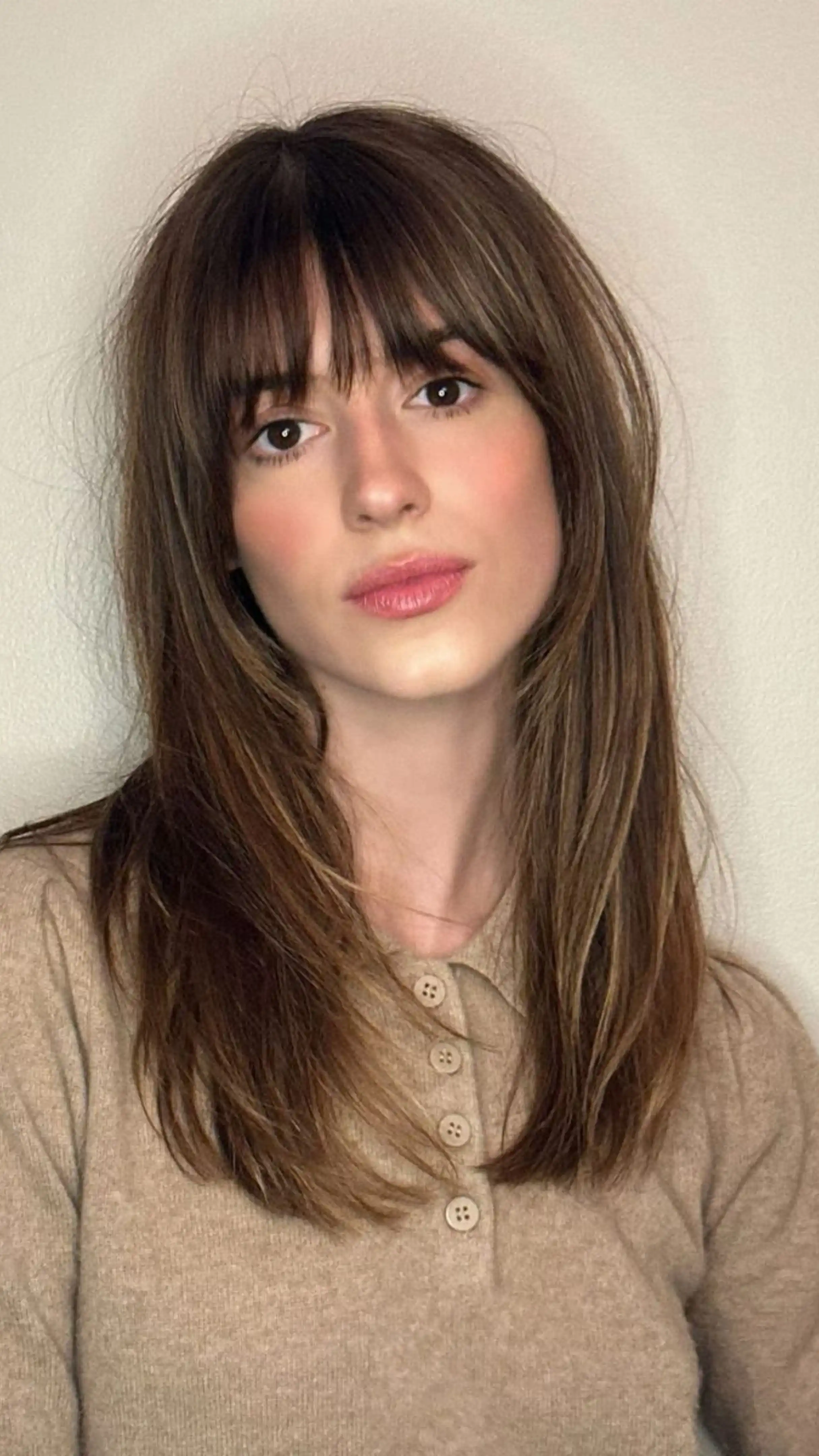 A woman with medium-length brown layered hair and birkin bangs, wearing a beige buttoned shirt, poses against a neutral background.