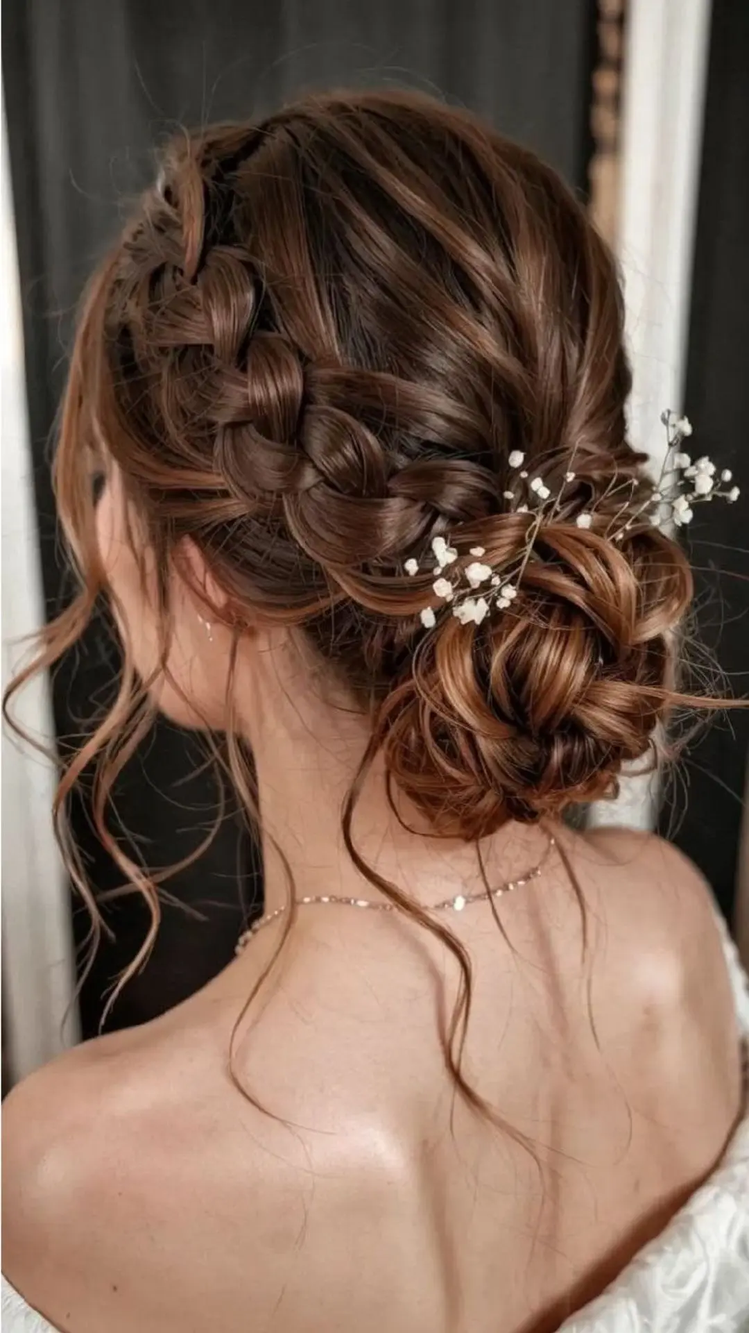 Rear view of a person with brown hair styled in an intricate side braid leading into a low, textured bun, adorned with delicate sprigs of baby's breath flowers.
