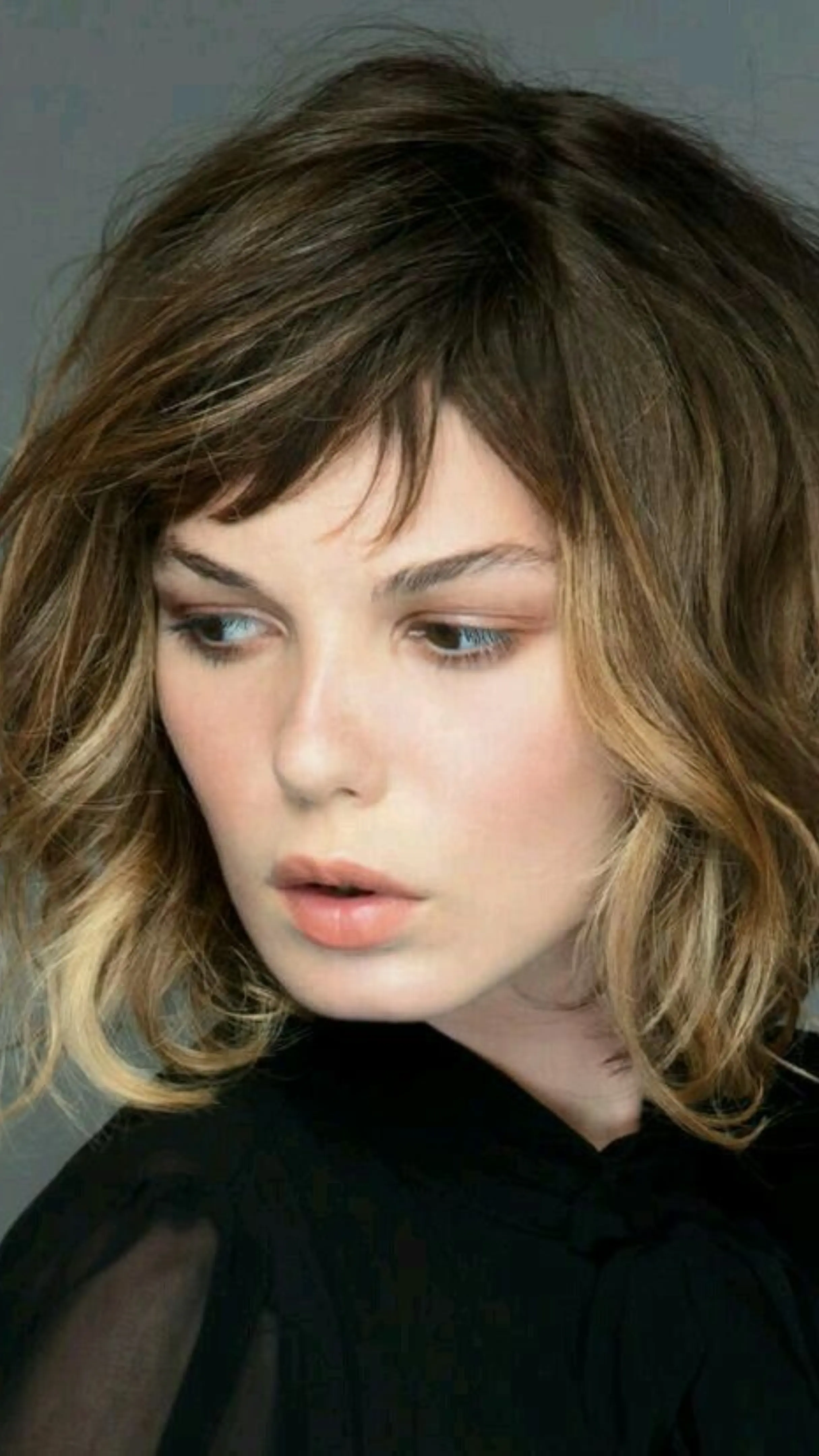 Close-up portrait of a woman with tousled, short brown and blonde ombre hair with choppy bangs, wearing a black top against a gray background.