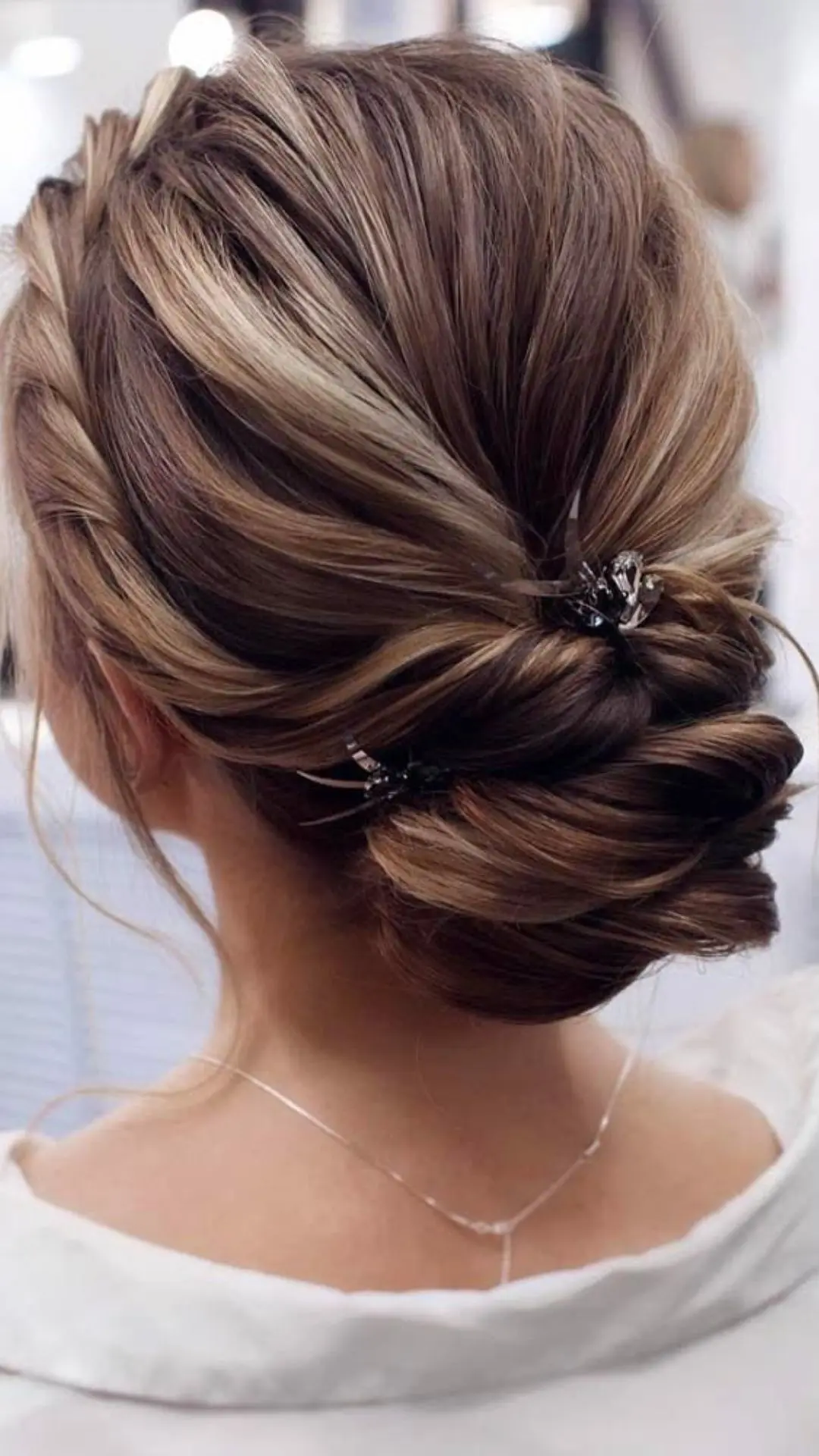 Back view of a woman with highlighted brown hair styled into an intricate, textured low updo with spider-shaped hairpins.