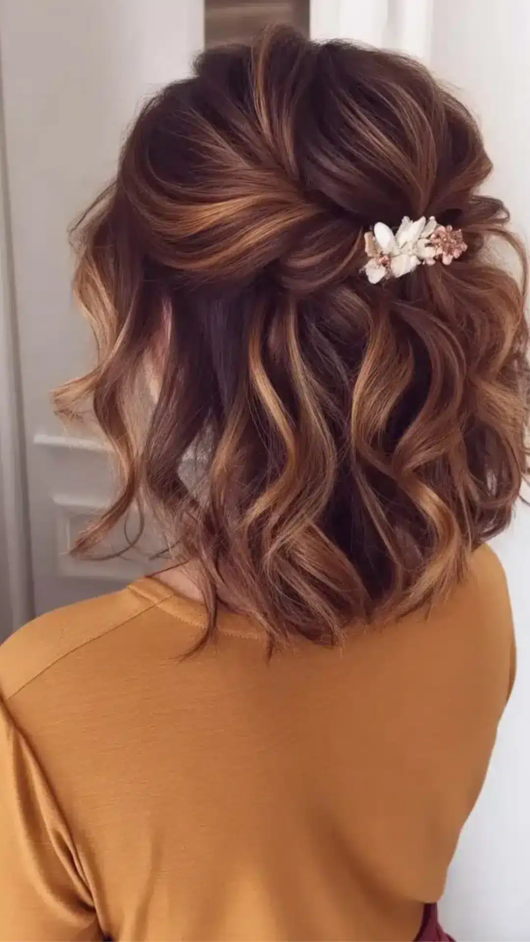 Back view of a person with shoulder-length, curly brown and highlighted hair styled in a half-up do secured with a white floral hair clip.