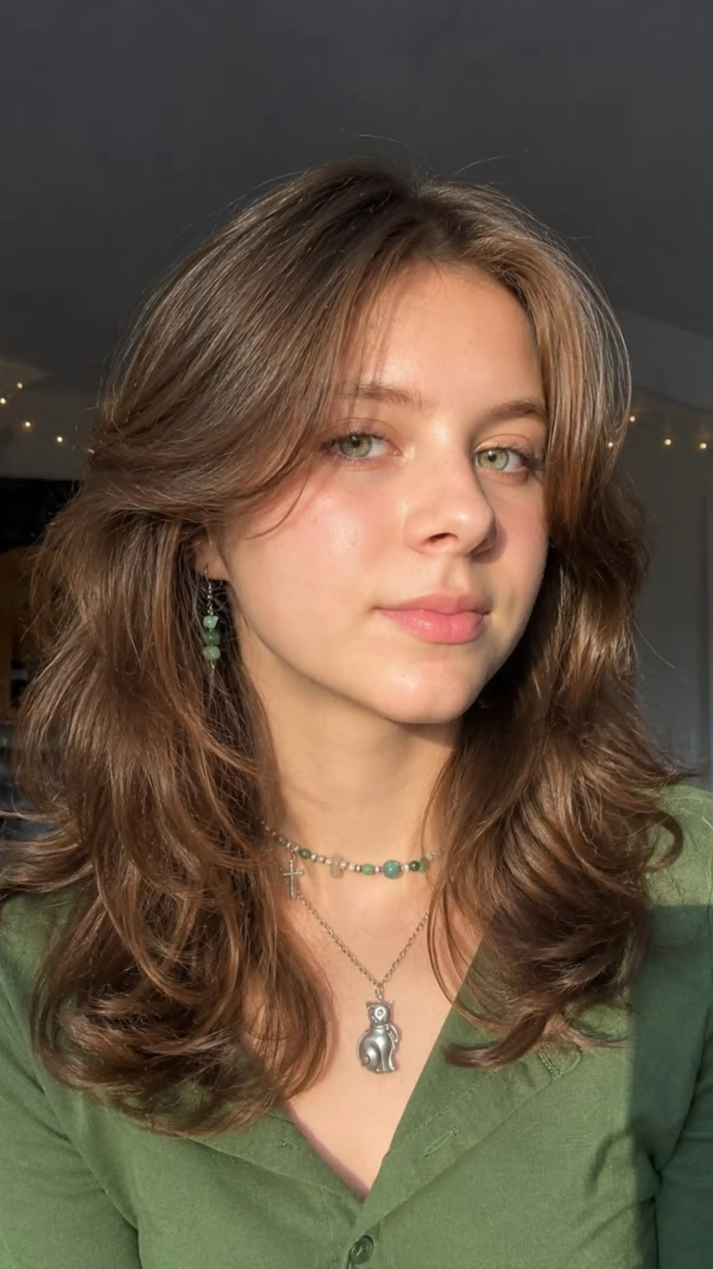 Close-up of a young woman with shoulder-length brown hair in layers with curtain bangs, green eyes, and a neutral expression, wearing a green top and layered necklaces with a cat pendant.