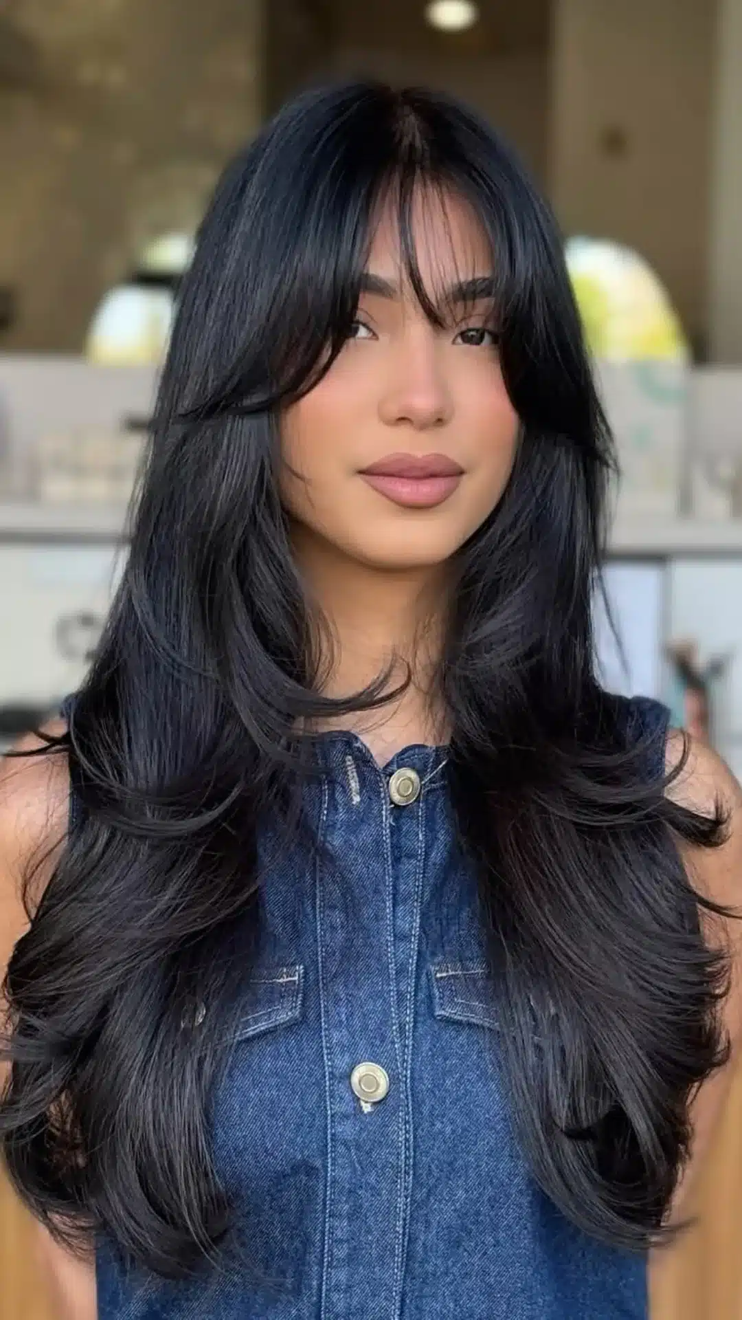 A young woman with long, black layered hair featuring dense bangs, wearing a dark blue denim top.