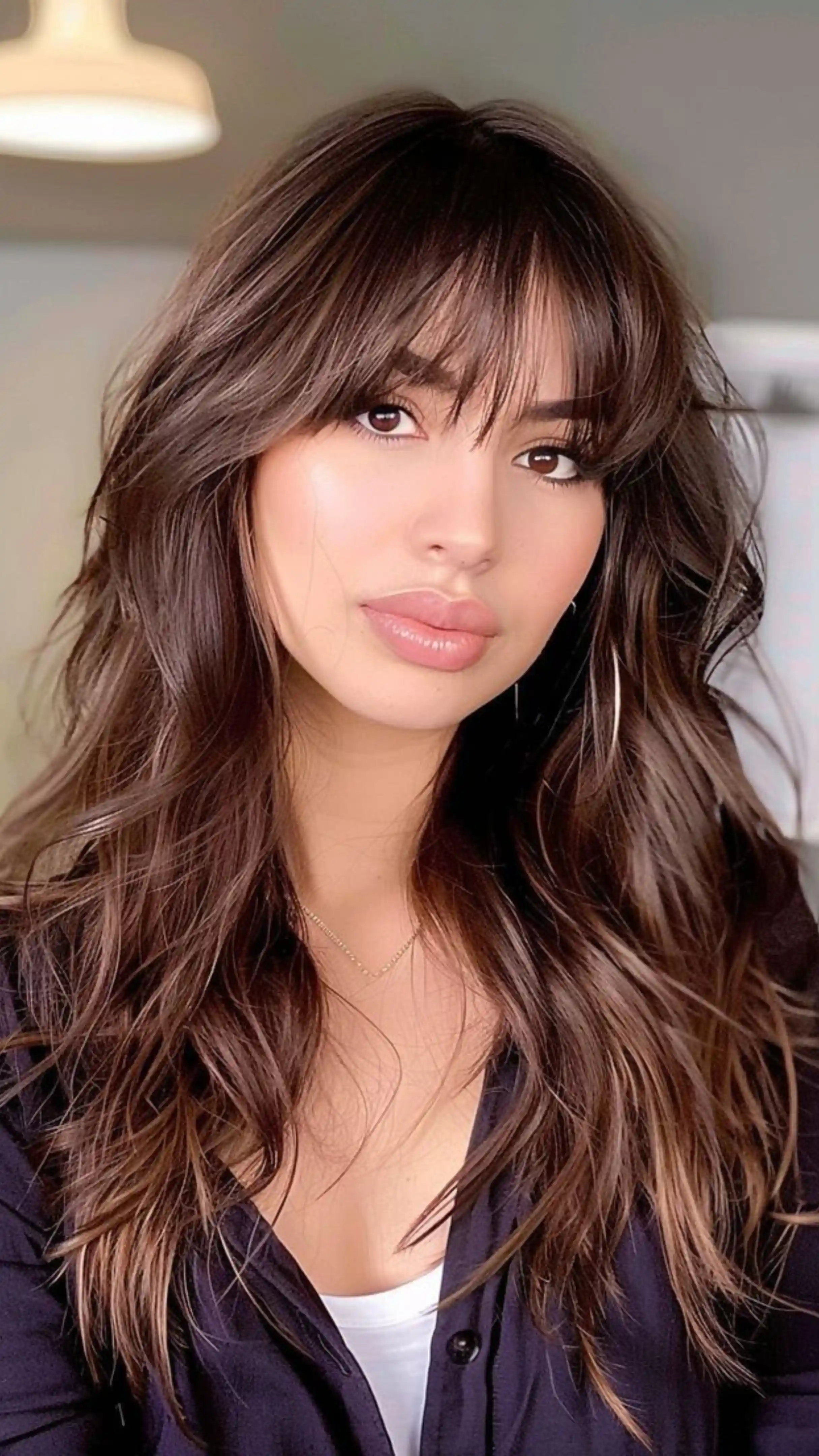 A young woman with long, layered brown hair featuring feathered bangs and soft waves looks toward the camera in a brightly lit indoor setting.