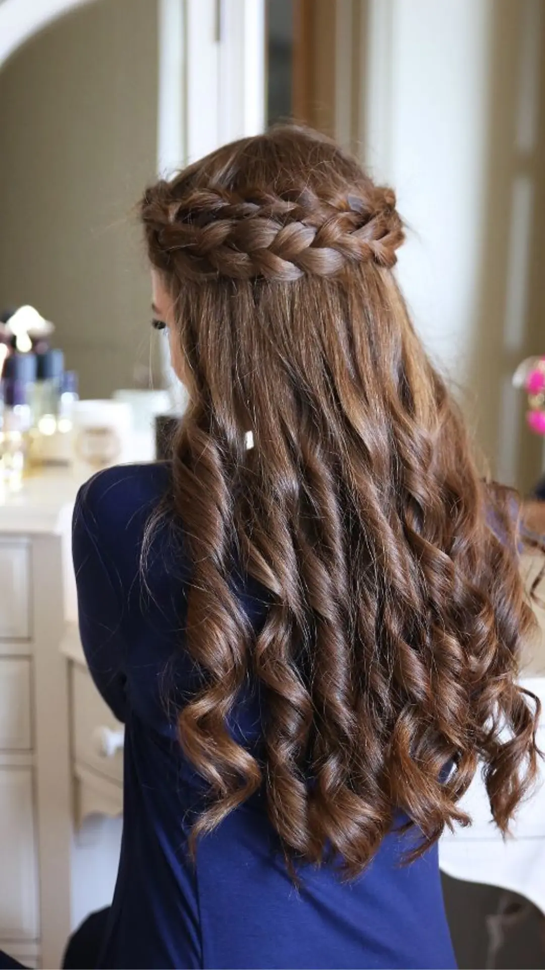 Back view of a person with long, brown hair styled with a crown braid on top and cascading curls below, wearing a blue shirt.