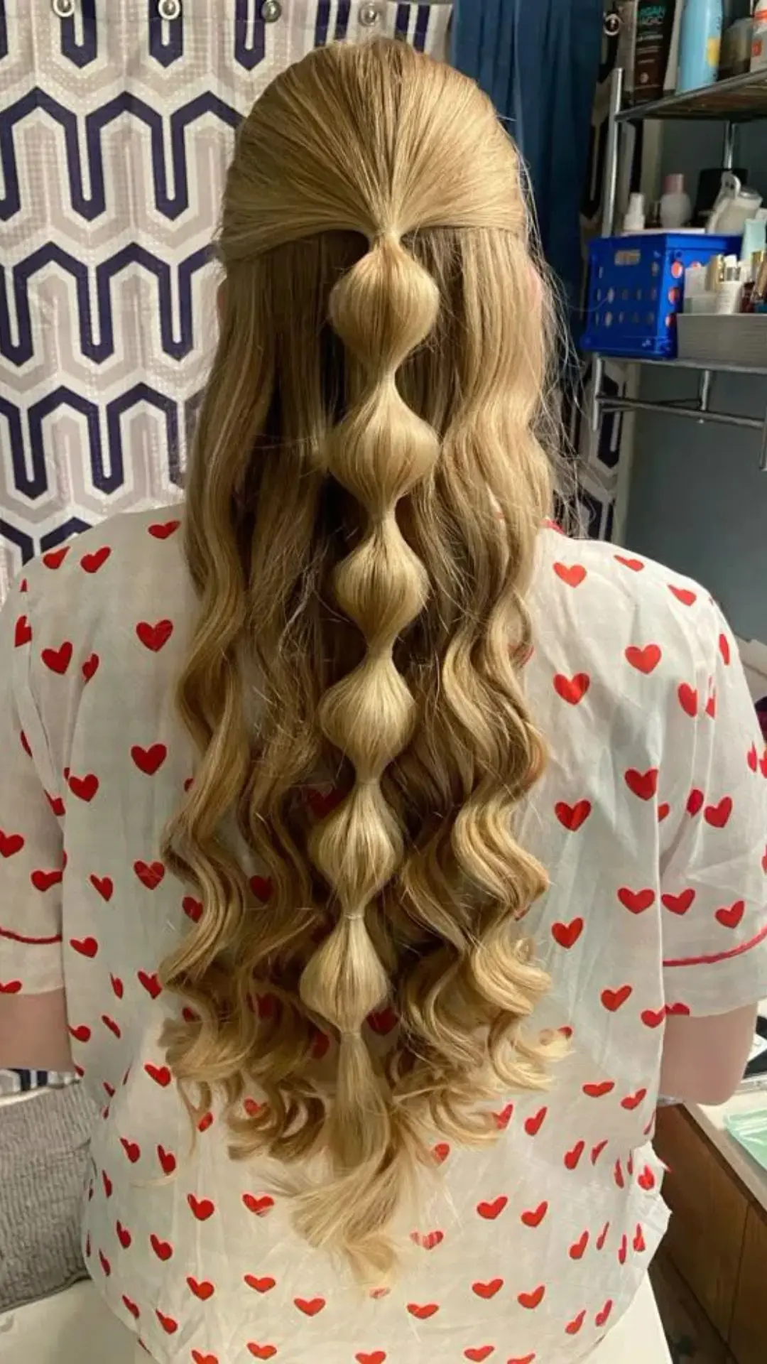 Back view of a person with long, blonde, wavy hair styled into a continuous bubble ponytail, wearing a white shirt with a red heart pattern.