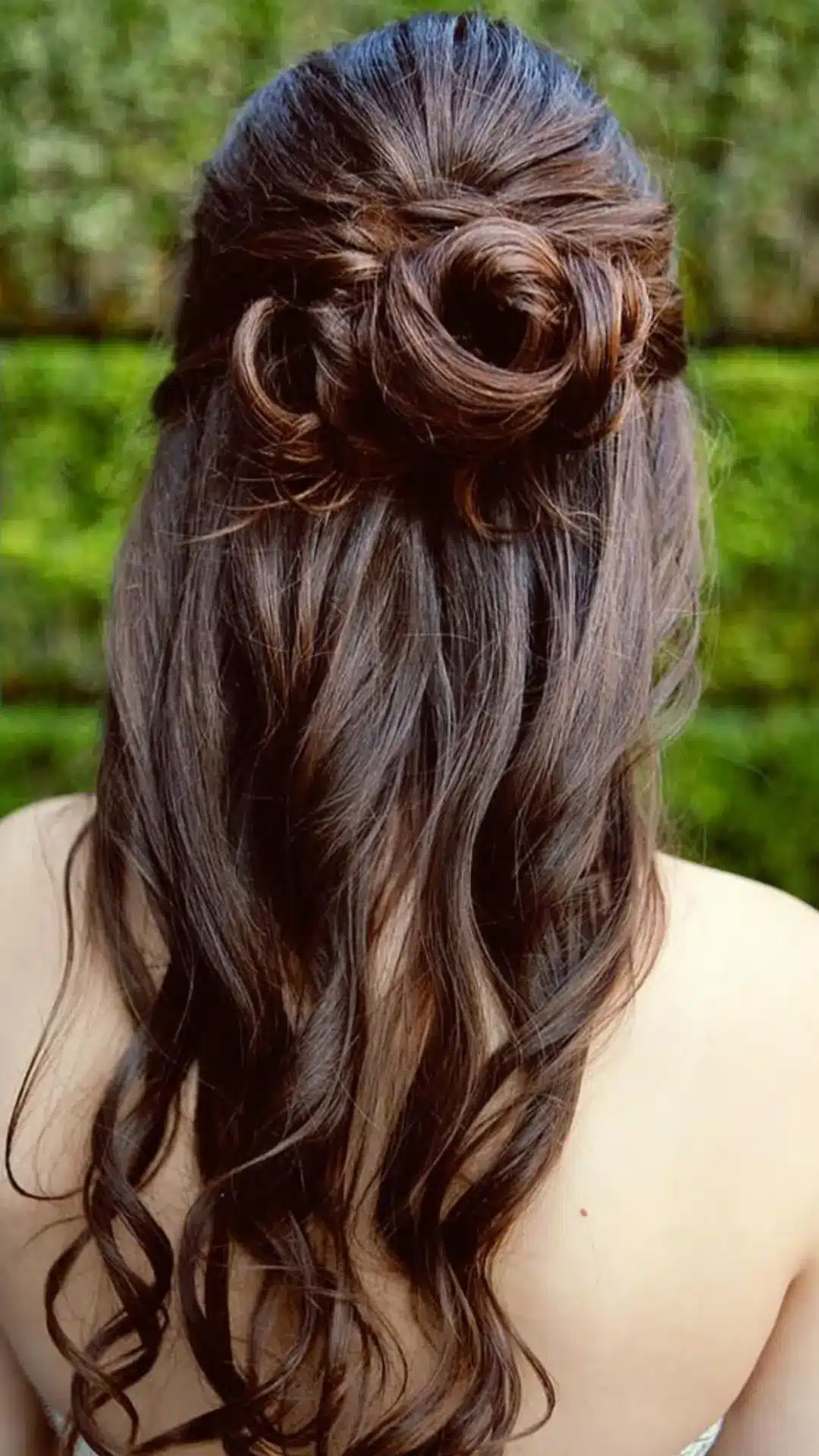 Back view of a person with long, dark brown hair styled in a half-up, half-down hairdo, featuring a twisted rose-like bun at the crown and loose, cascading waves falling down the back, set against a blurred green background.