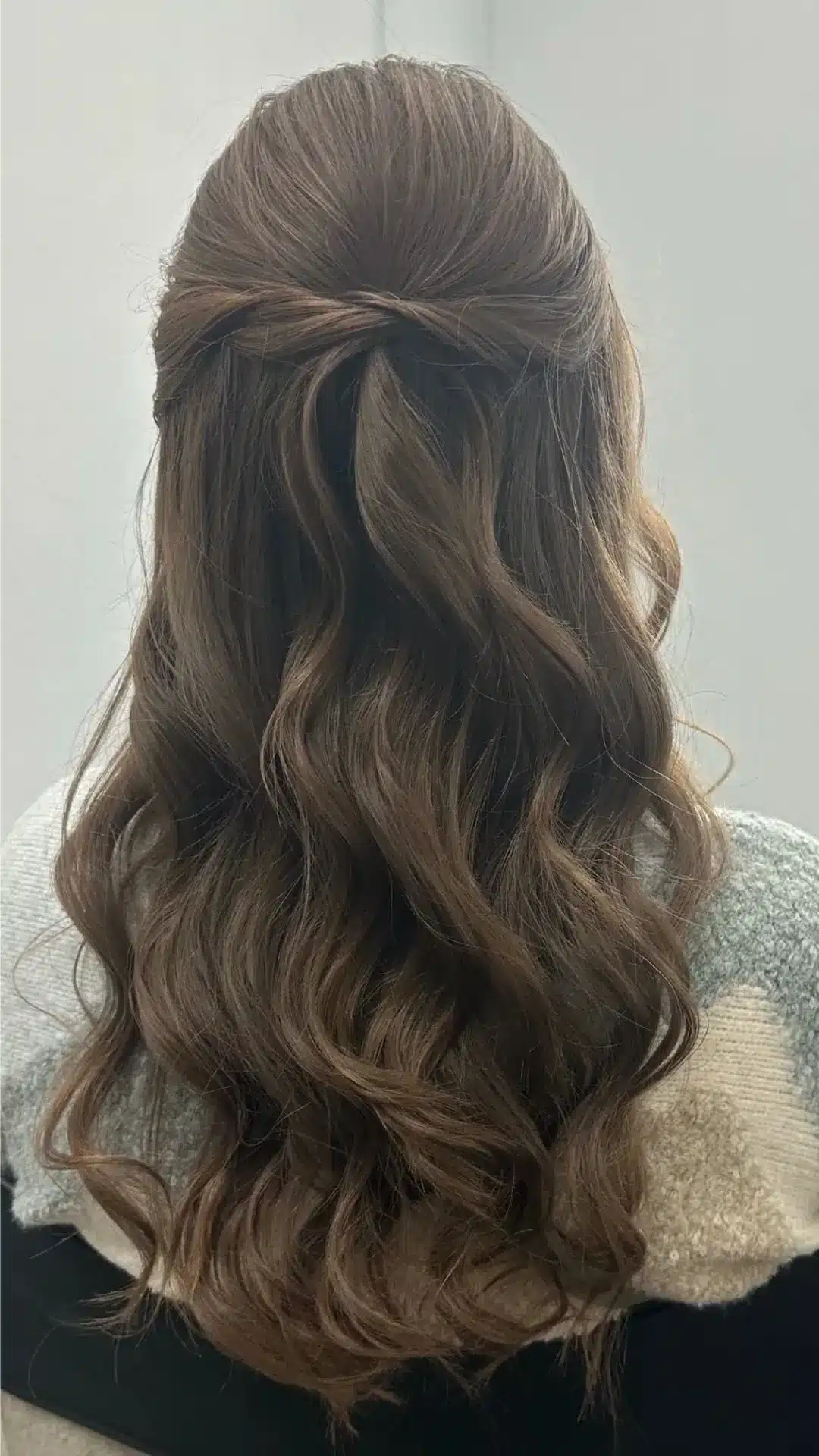 Rear view of a person with long, wavy brown hair styled in a half-up twist, wearing a gray and white textured sweater against a plain background.
