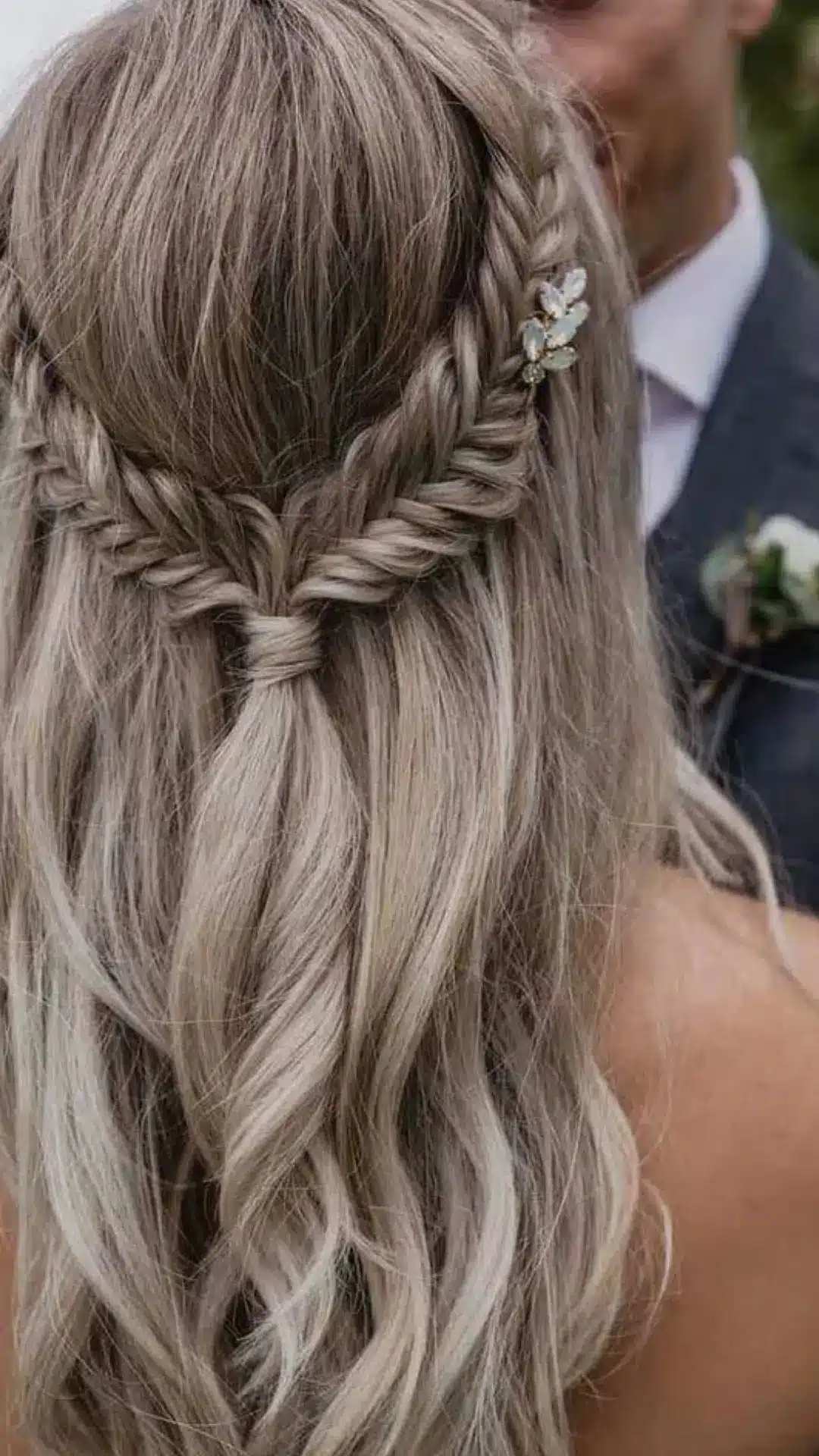 Back view of a bride with long, ash-blonde, wavy hair styled in a half-up fishtail braid secured with a hairpiece, standing next to a groom in a dark suit.