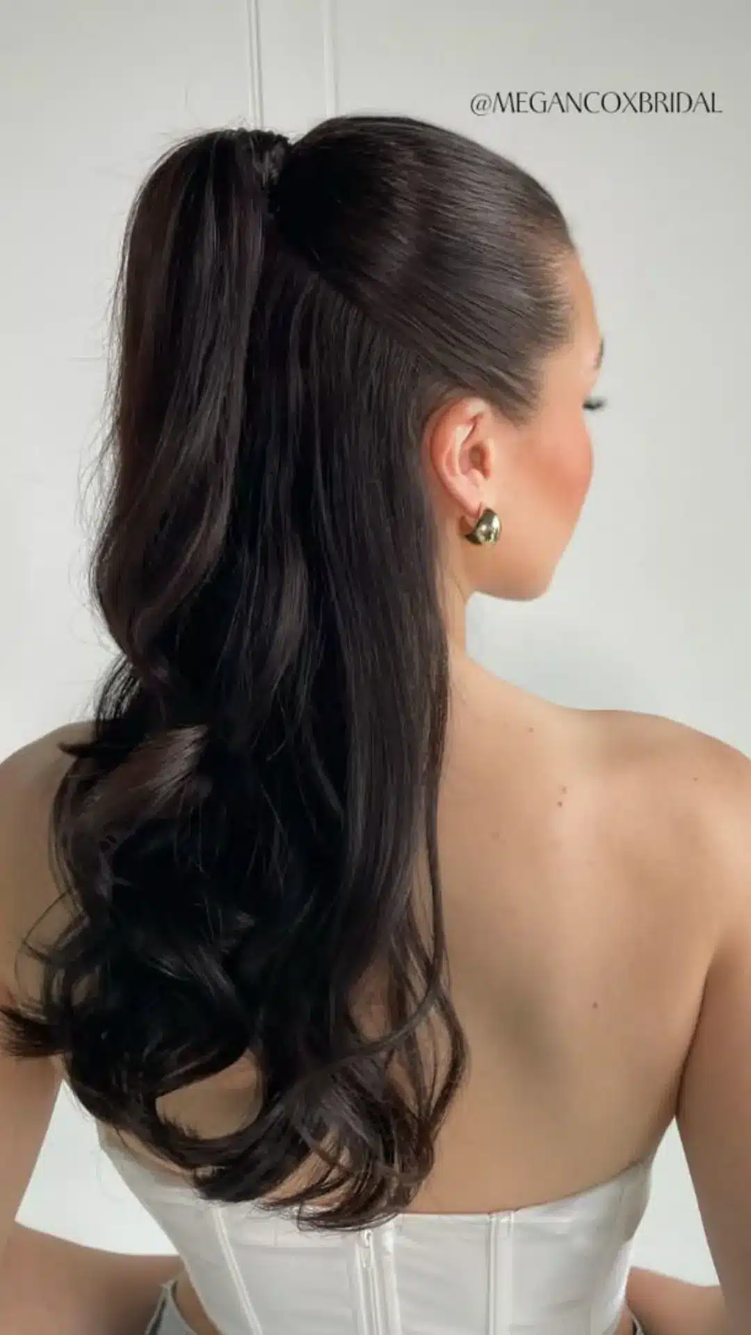 Back view of a person with dark, long, voluminous hair styled in a half-up, half-down ponytail with soft curls at the ends, wearing a strapless white top and gold hoop earrings.