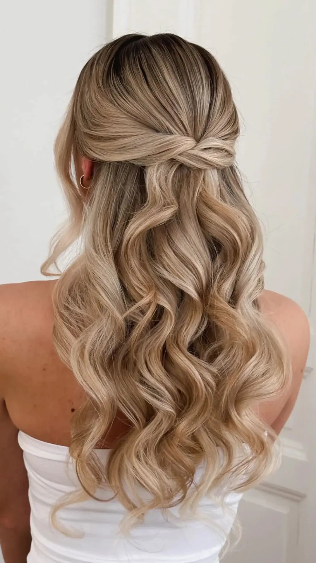 Rear view of a person with blonde, highlighted hair styled in a half-up, half-down twist with voluminous curls cascading down their back.