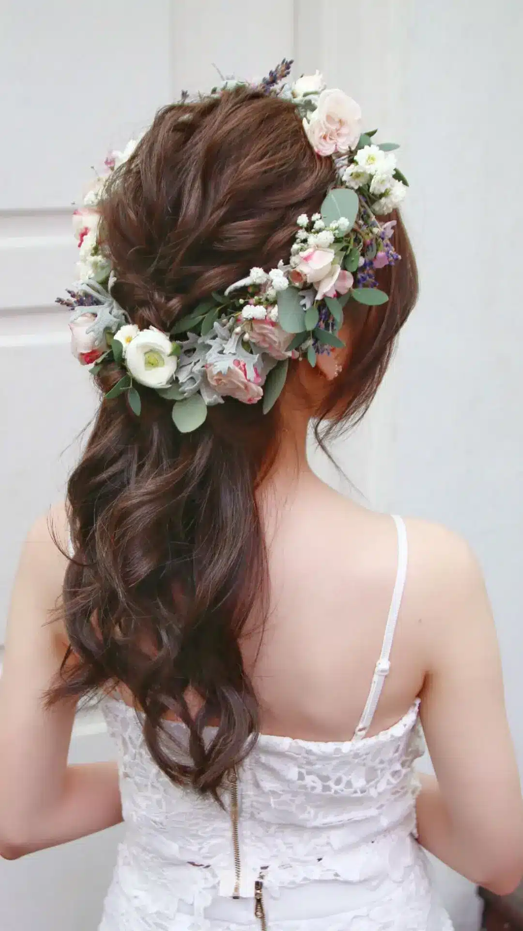 Rear view of a woman with long, dark, wavy hair styled half-up and adorned with a lush floral crown of pink and white roses, greenery, and small white flowers, wearing a white lace dress.