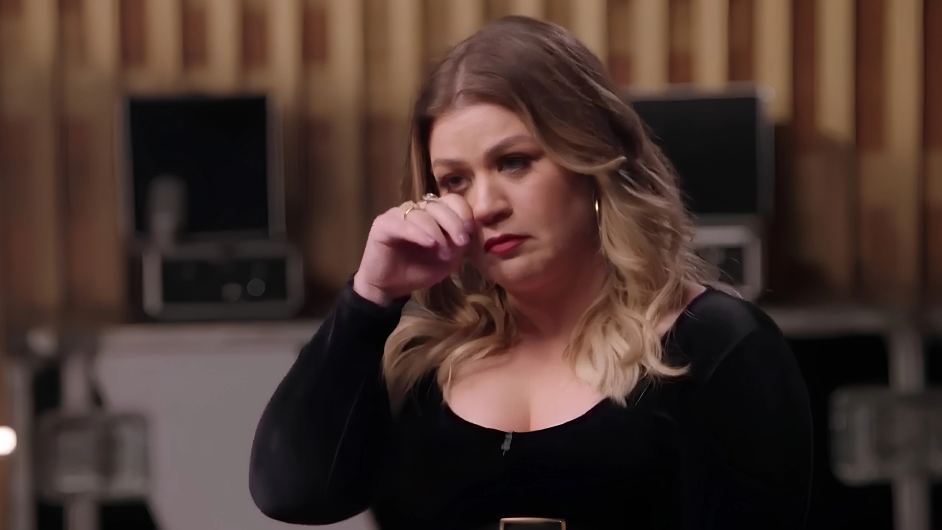 Kelly Clarkson dabs her eye with her hand while wearing a black dress