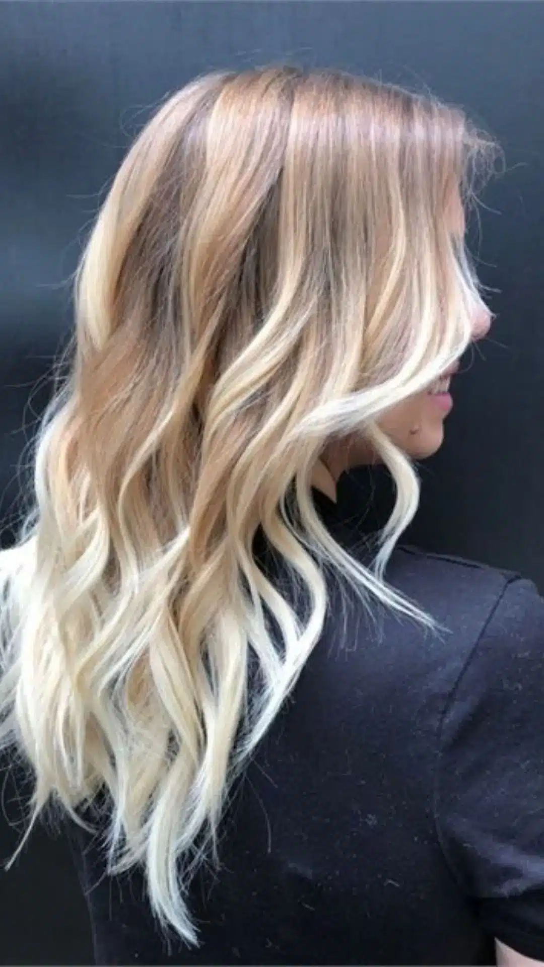 Profile view of a woman with long, wavy blonde ombre hair featuring darker roots transitioning to platinum blonde ends, wearing a black shirt against a dark background.