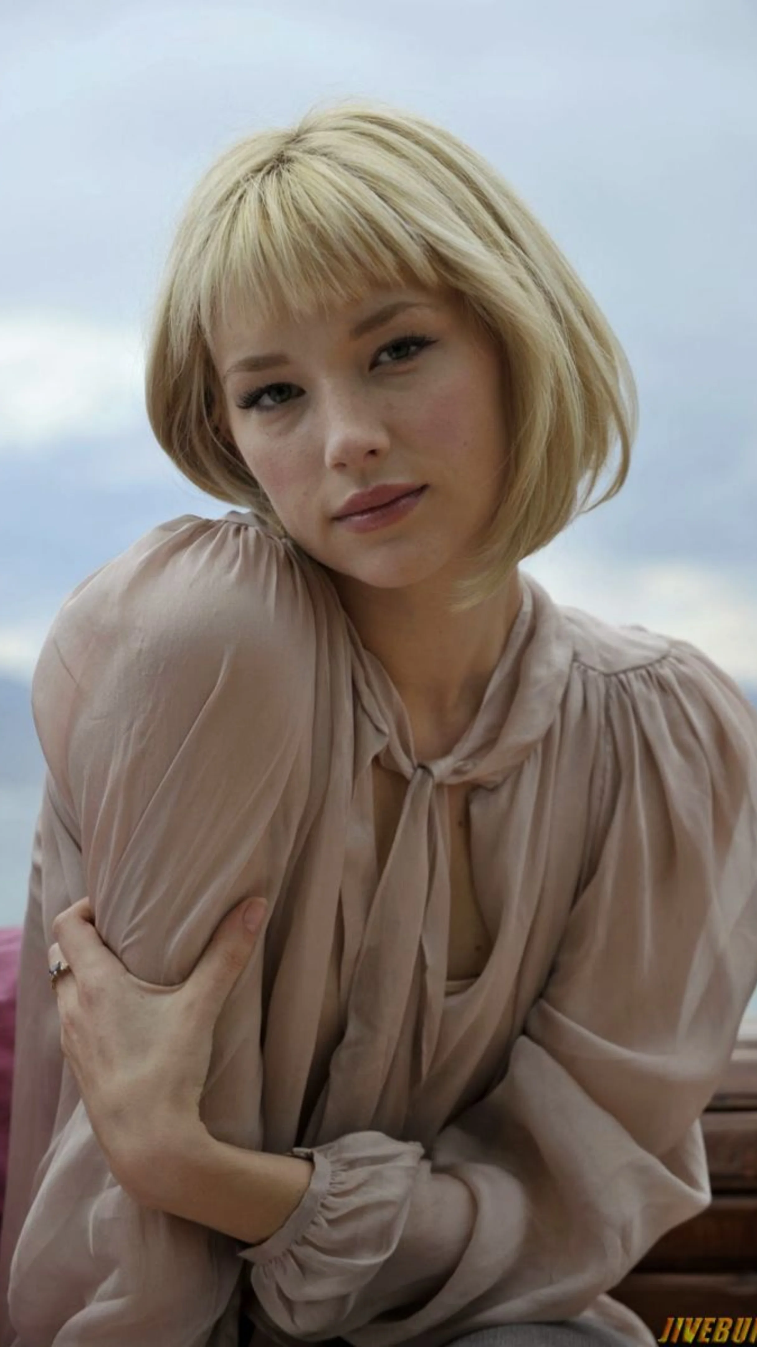 A blonde woman with short hair and micro bangs wears a long-sleeve, silky light pink top with a tie at the collar while looking thoughtfully at the camera.
