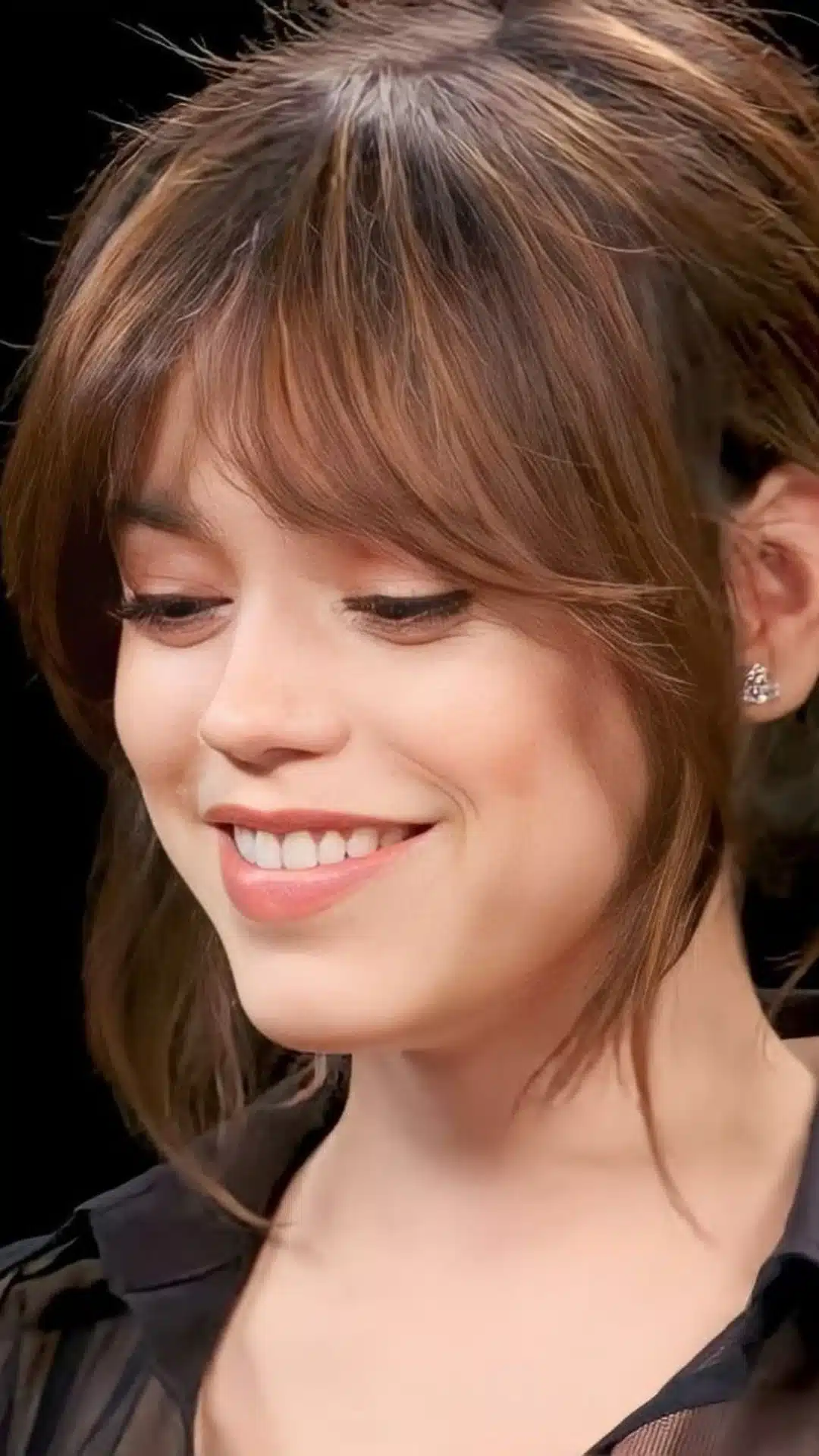 Close-up of a smiling young woman with brown hair and piecey bangs against a black background.