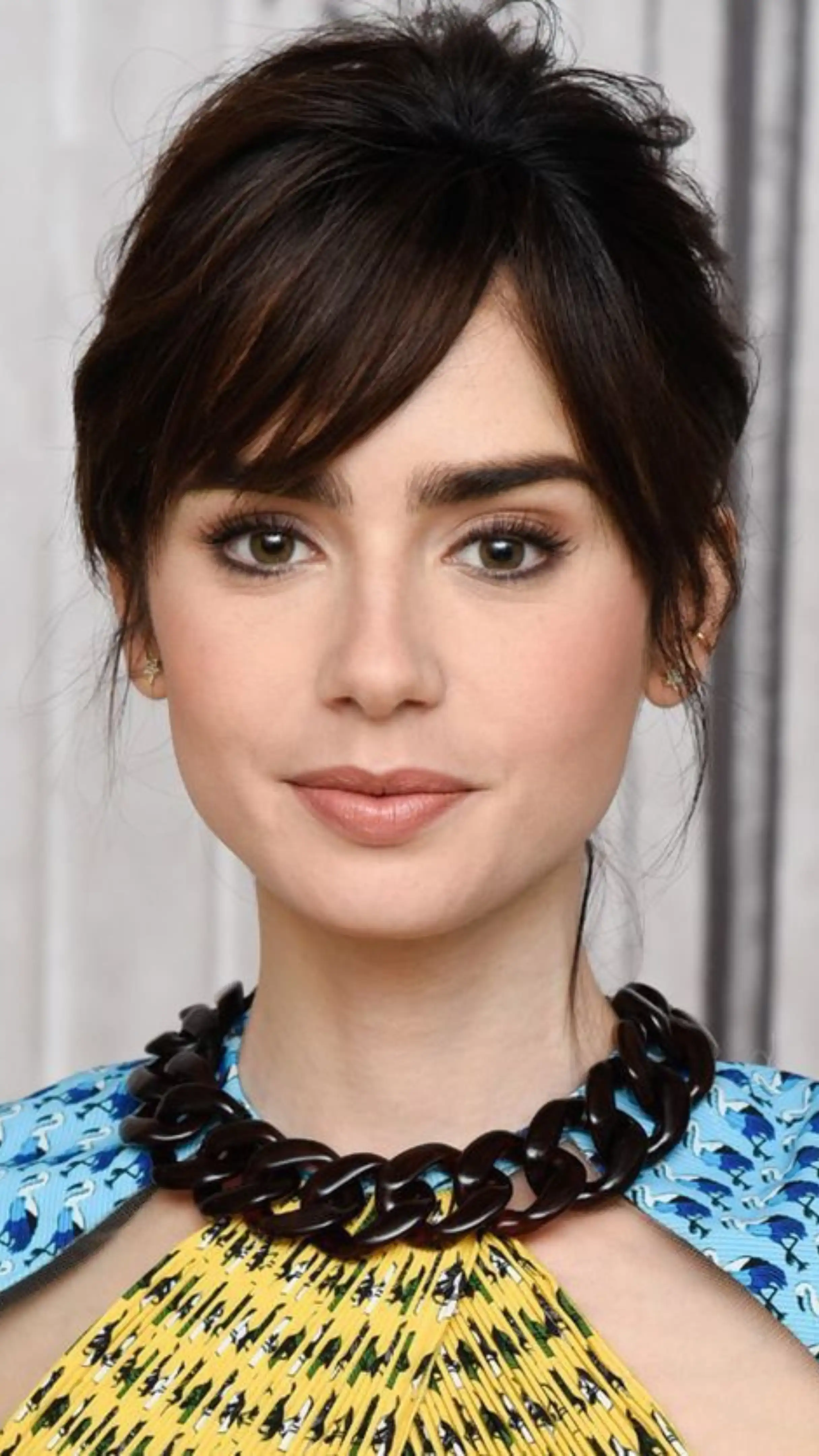 Close-up portrait of actress Lily Collins with dark hair, side- swept bangs, and a bold black chain necklace against a blurred background.