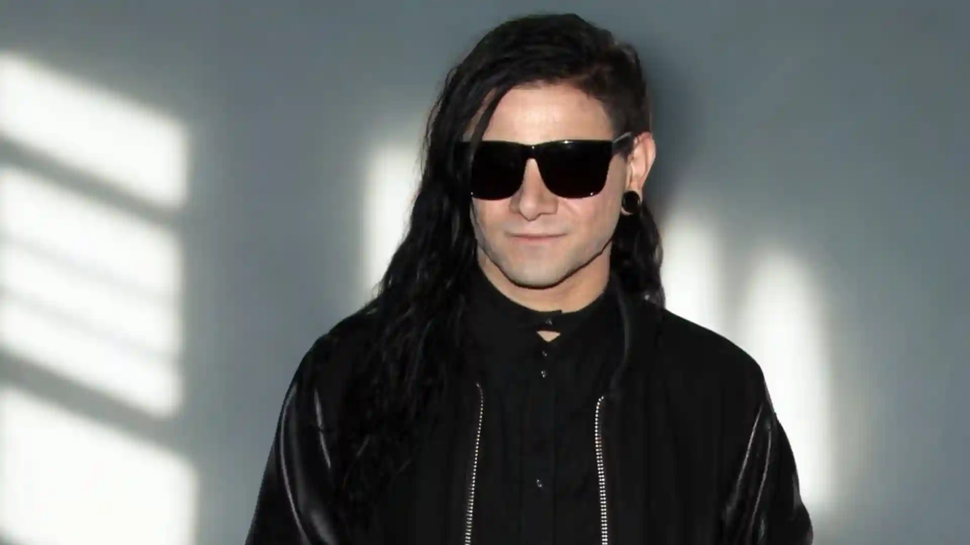 Skrillex wearing sunglasses and a black outfit stands against a gray wall with light patterns.
