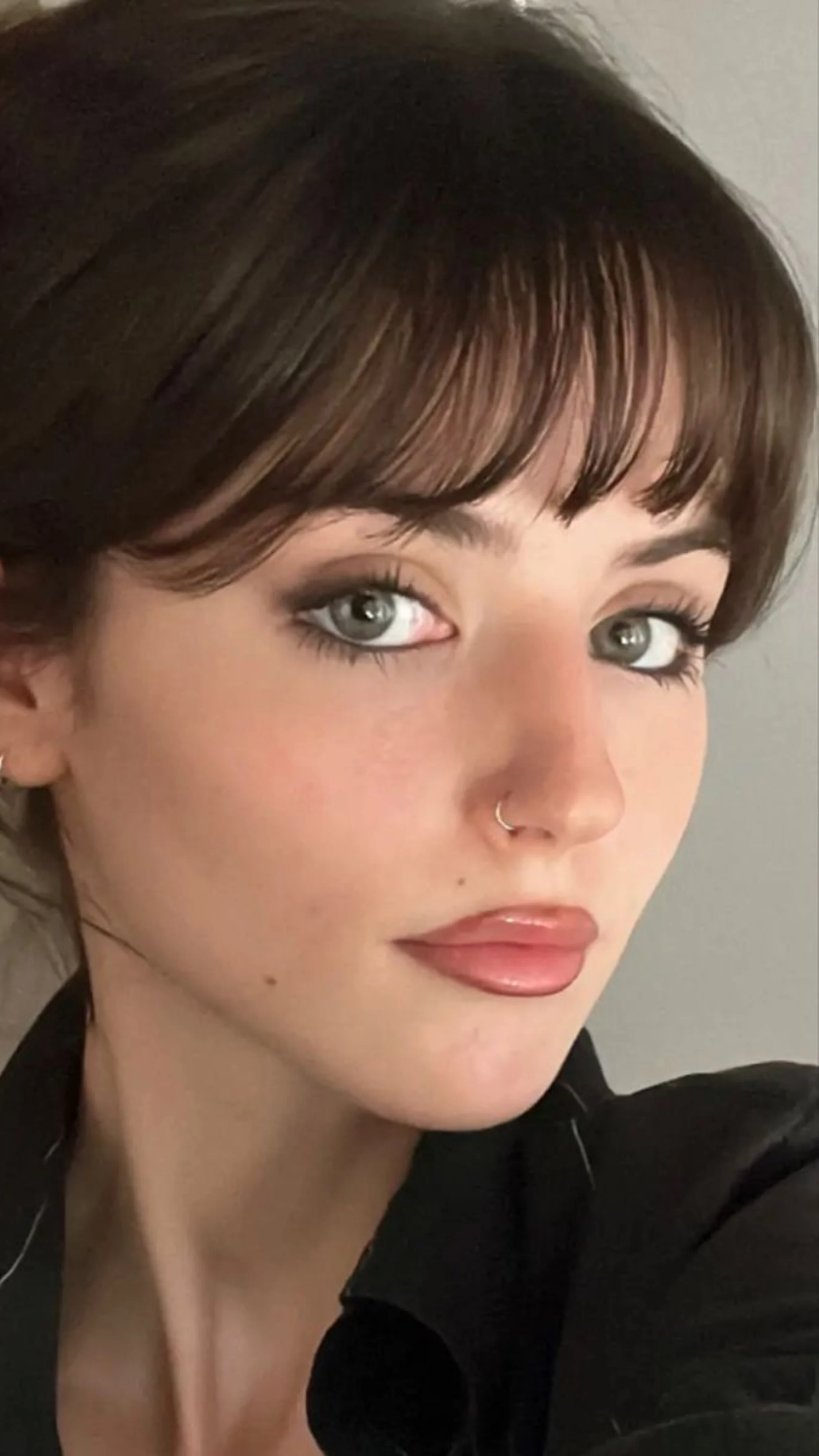 Close-up portrait of a woman with dark hair, soft half-moon fringe, and light eyes, wearing makeup with dark lip liner and a small nose ring.