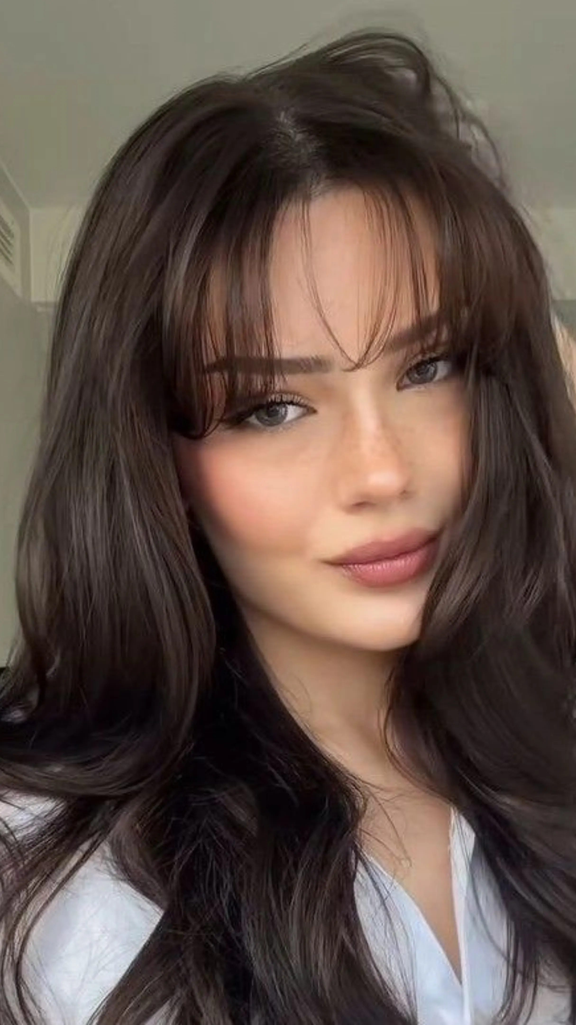 Close-up portrait of a woman with long, dark brown hair styled with wispy bangs, wearing light makeup and looking directly at the camera with a subtle smile.