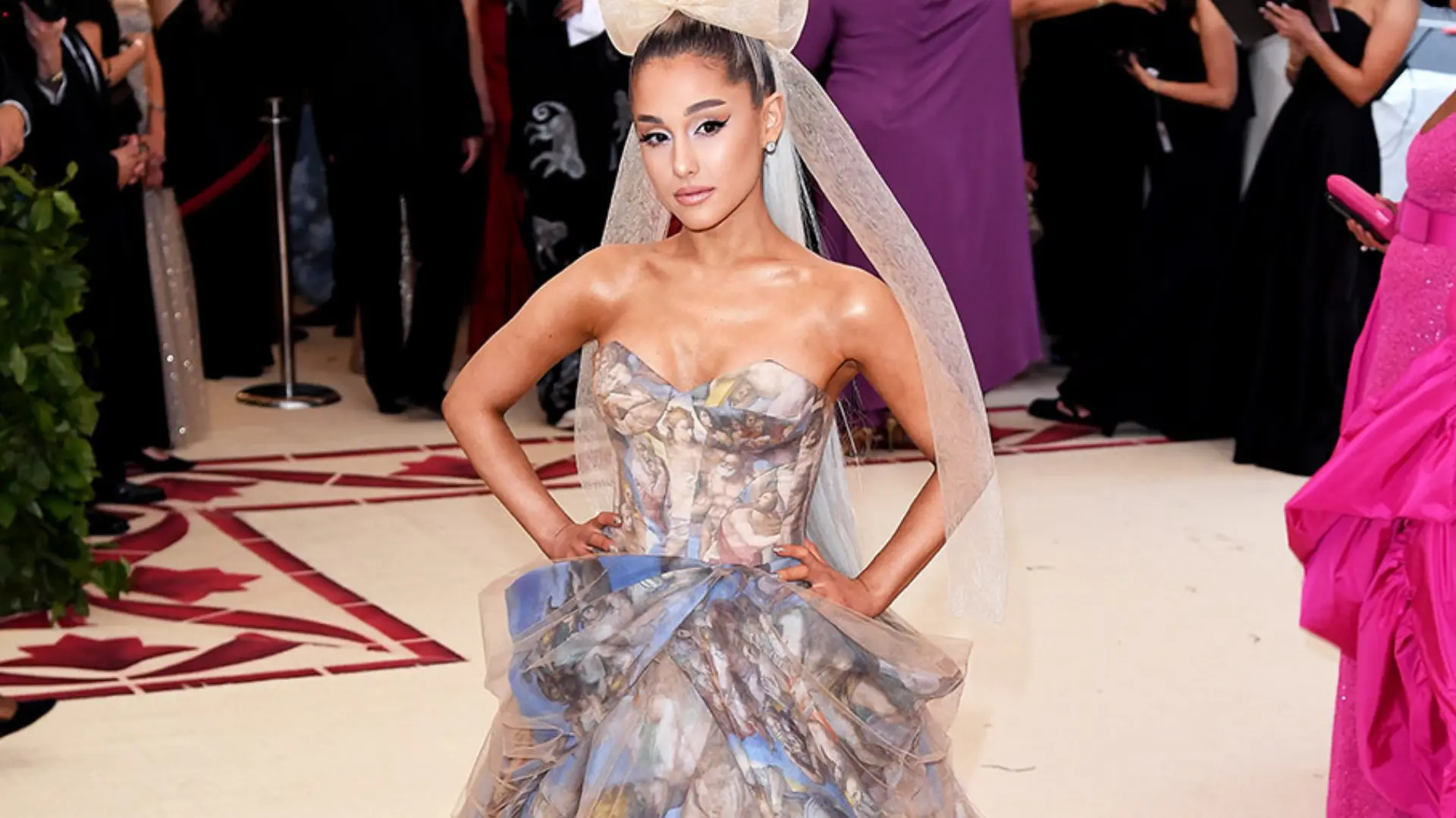Ariana Grande poses on a red carpet in a strapless patterned gown with a large bow and flowing veil.