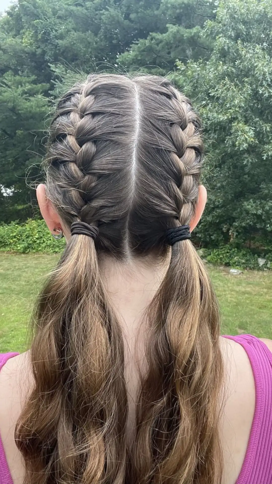 back view of french braid pigtails on long wavy hair with center part simple braided hairstyle in natural outdoor setting