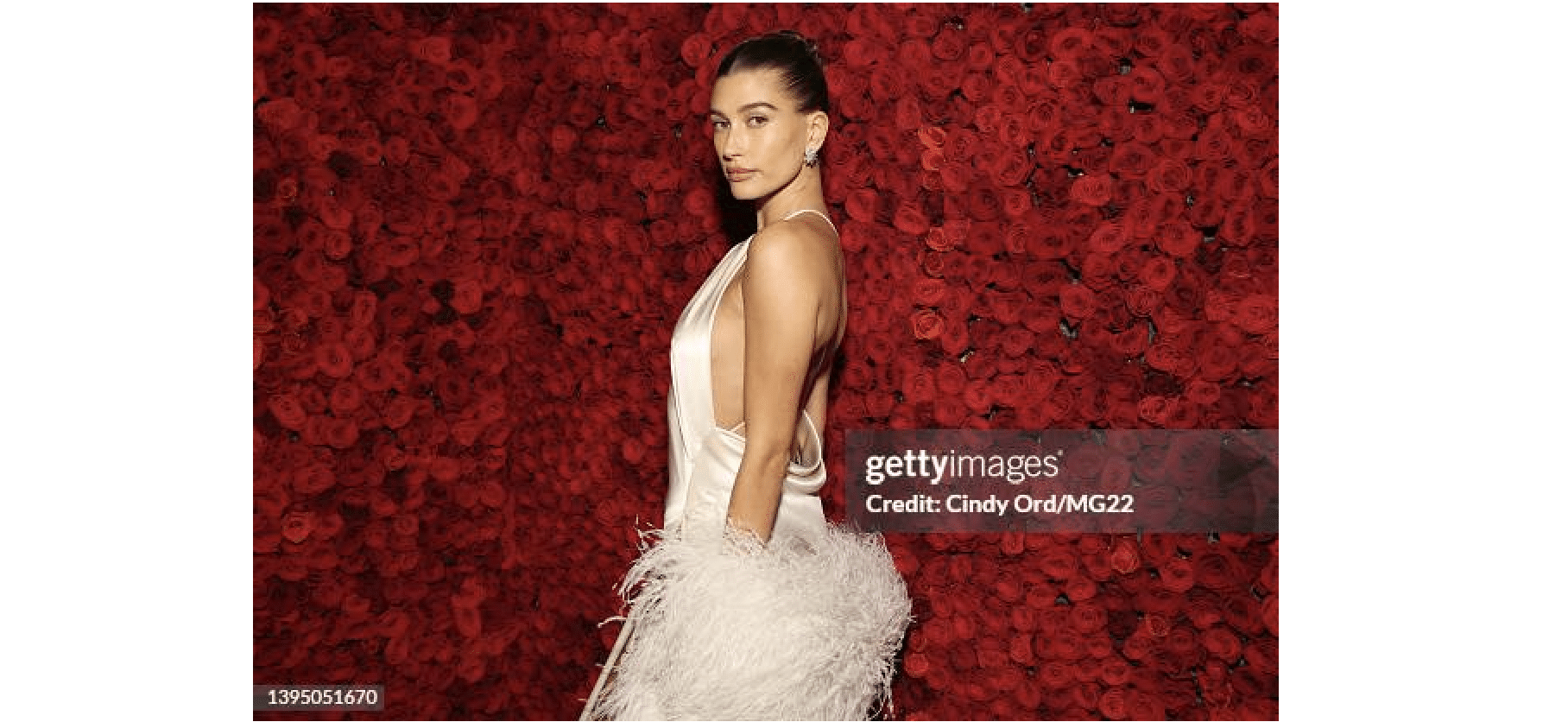 Hailey Bieber in a sleek bun posing in a satin gown before a wall of red roses at Met Gala 2022