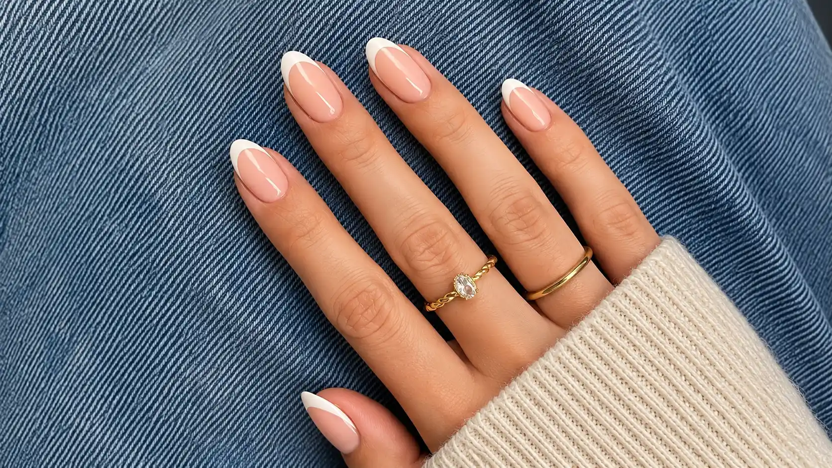 Glossy almond french tip nails with delicate gold rings resting on textured blue denim beside an ivory cuff