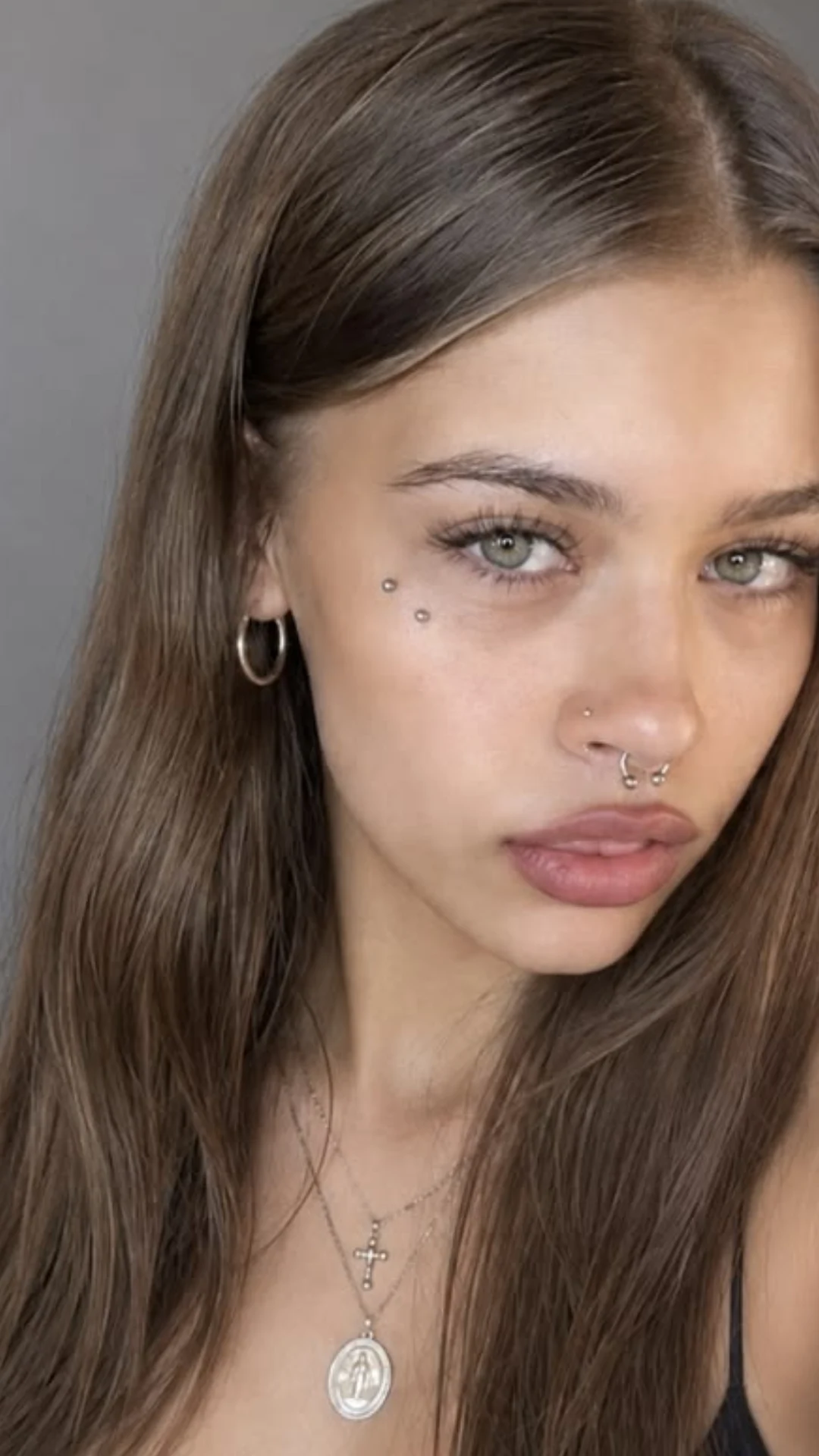 Close-up portrait of a young woman with long brown hair, green eyes, facial piercings, hoop earrings, and layered necklaces.