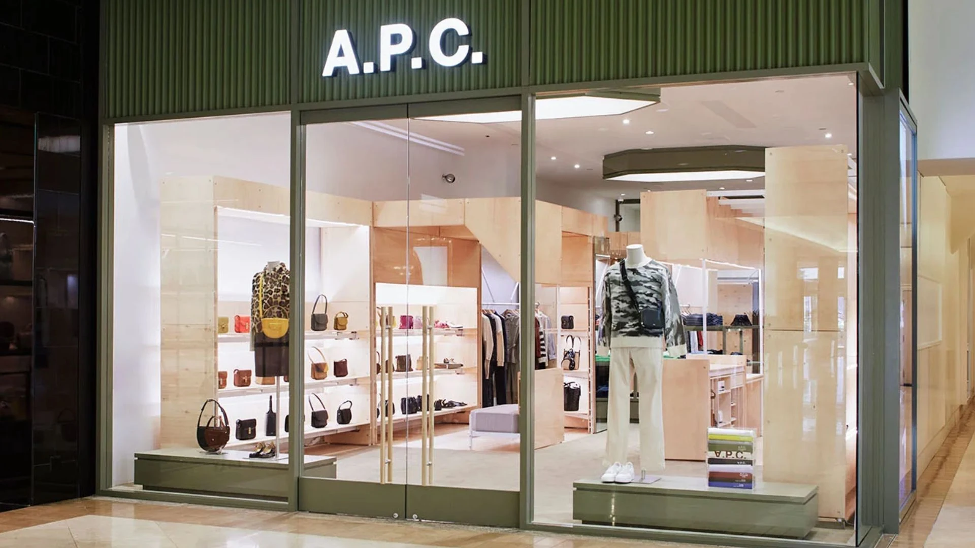 A.P.C. fashion boutique storefront displaying handbags, clothing, shoes, and accessories behind glass doors in a shopping mall.