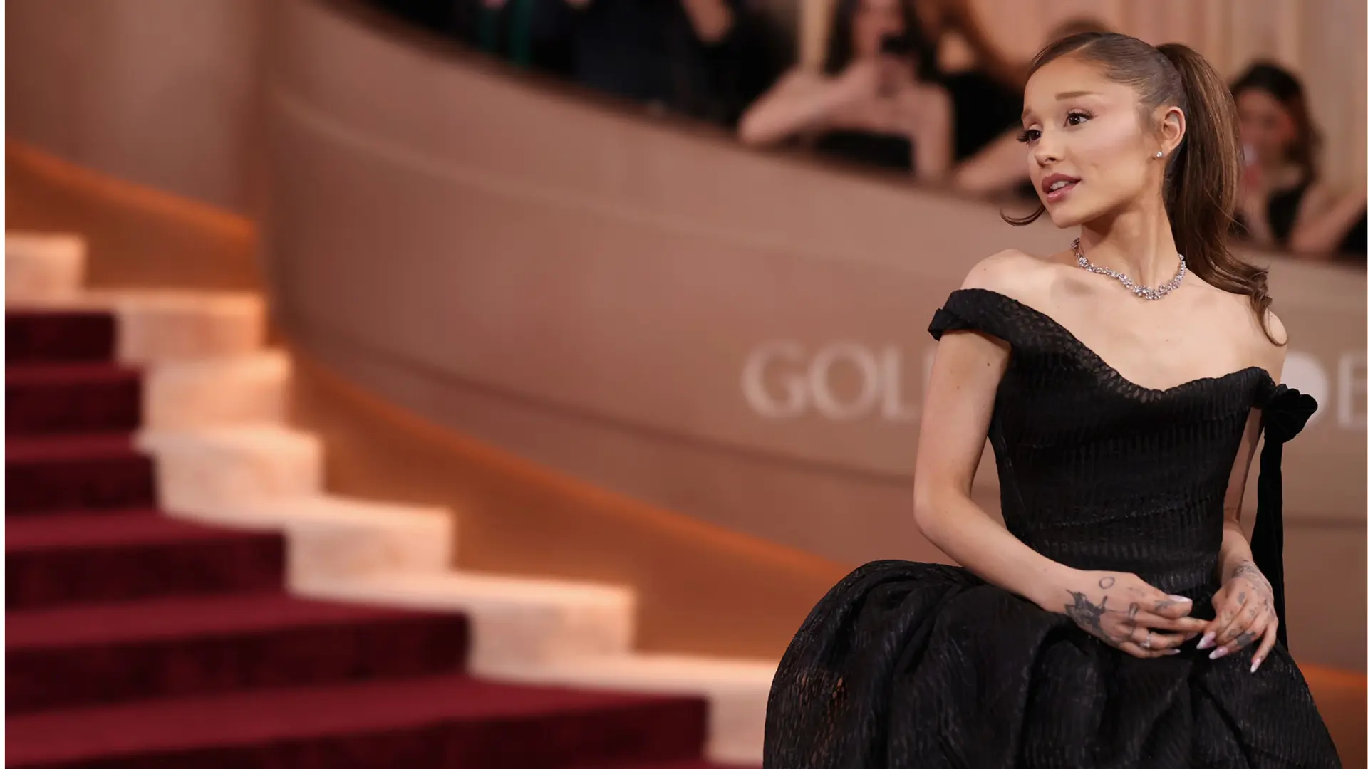 Ariana Grande poses in a black off-the-shoulder gown beside a red carpet staircase at a glamorous event.