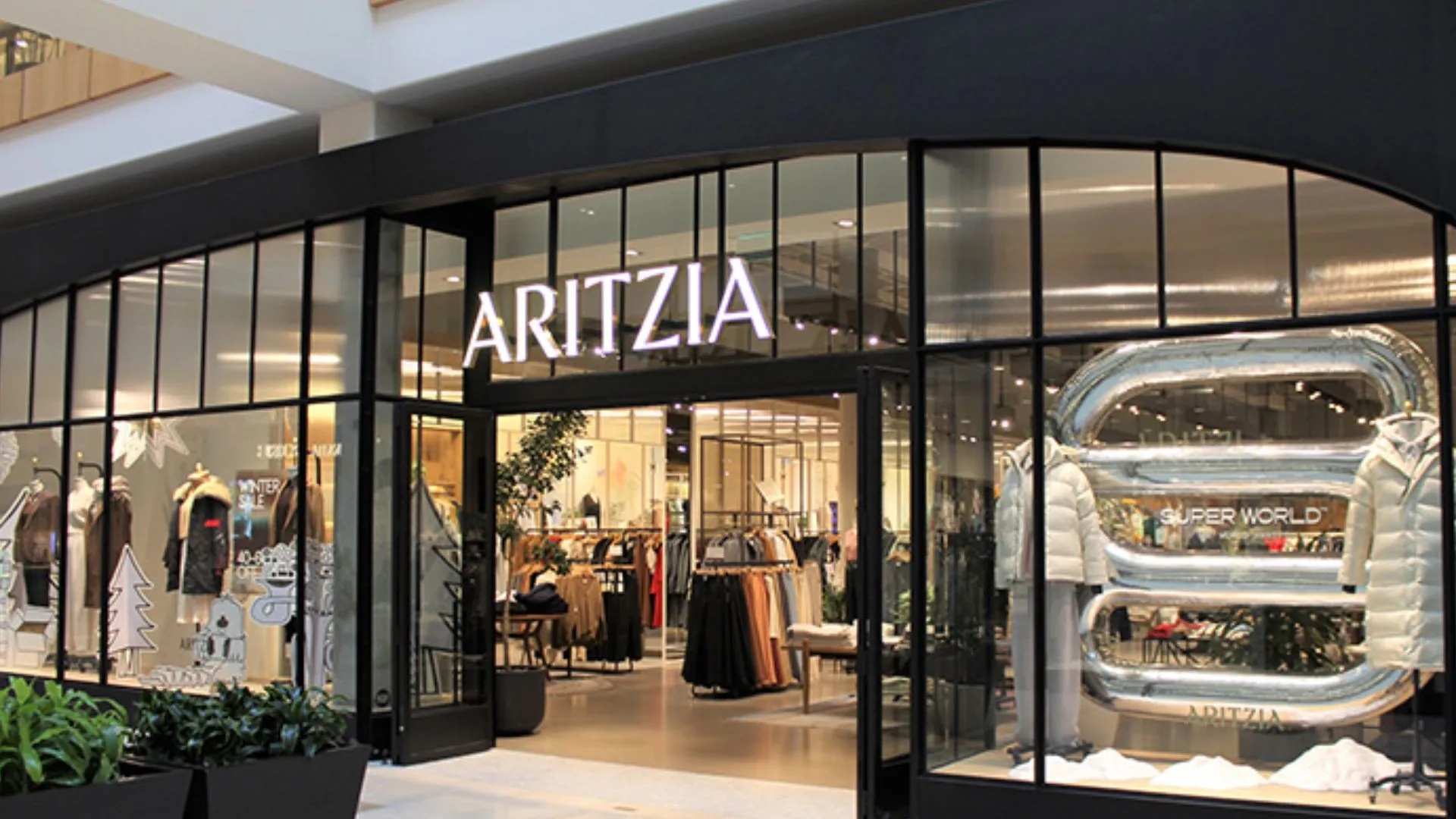Aritzia clothing store storefront in a shopping mall, featuring large glass windows, illuminated signage, clothing racks, and a silver “Super World” display.