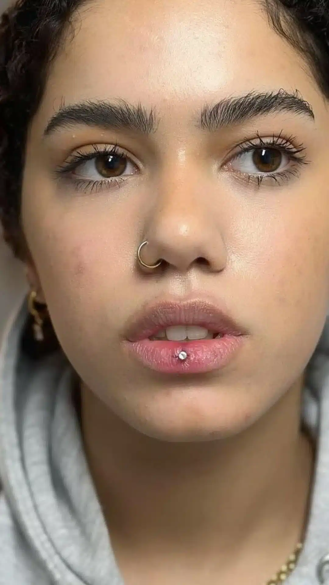 Close-up portrait of a woman with a gold nose ring, natural makeup, and a silver Ashley piercing centered on her lower lip.