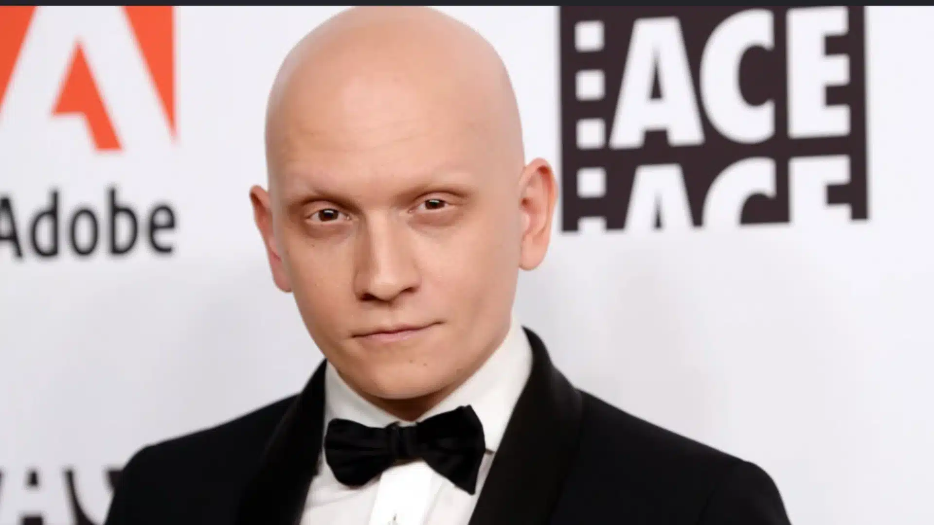 bald man in a black tuxedo and bow tie poses at a formal event against a branded black and white media backdrop