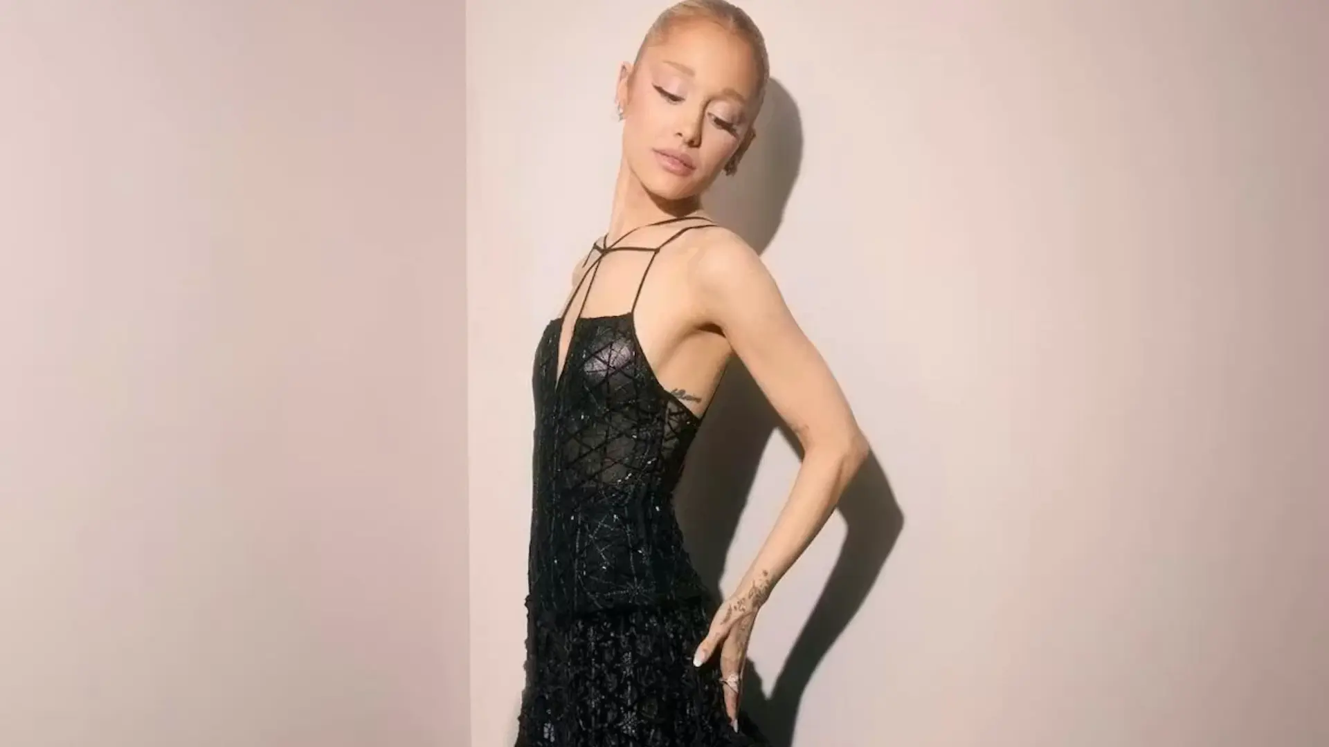 Model with a sleek blonde bun poses in a black lace dress against a pale pink wall, casting a dramatic shadow.