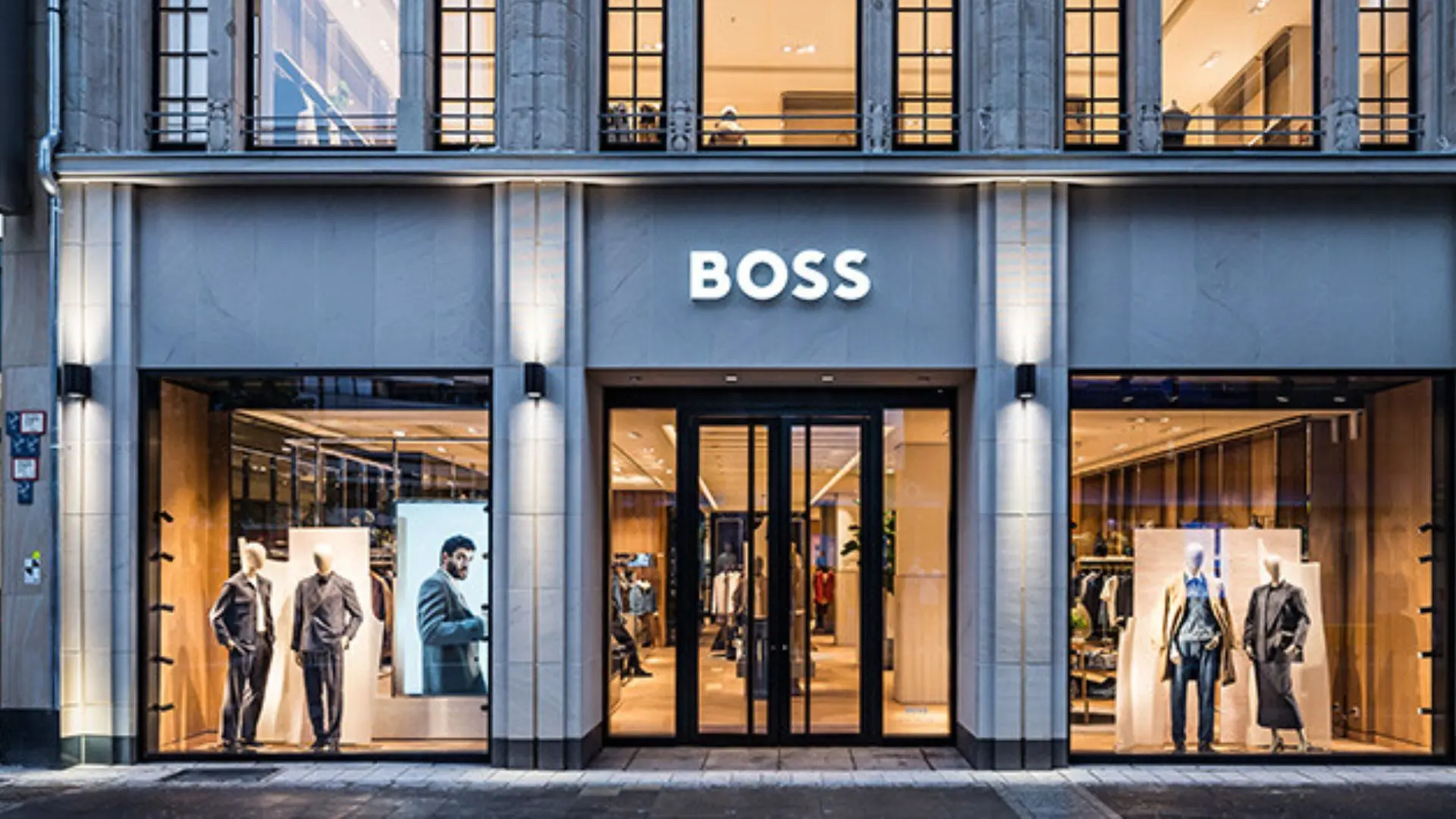 BOSS clothing store storefront with illuminated signage, glass entrance, and mannequins displaying men’s fashion in the window displays.