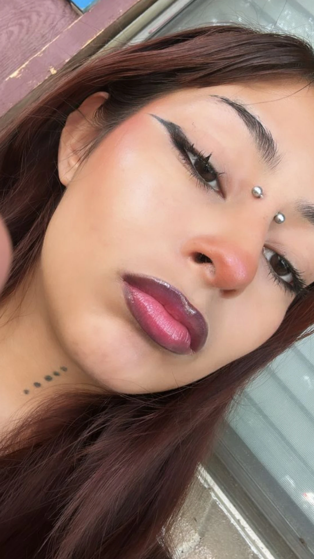Close-up portrait of a woman with winged eyeliner, glossy dark mauve lipstick, forehead piercings, and long auburn hair.