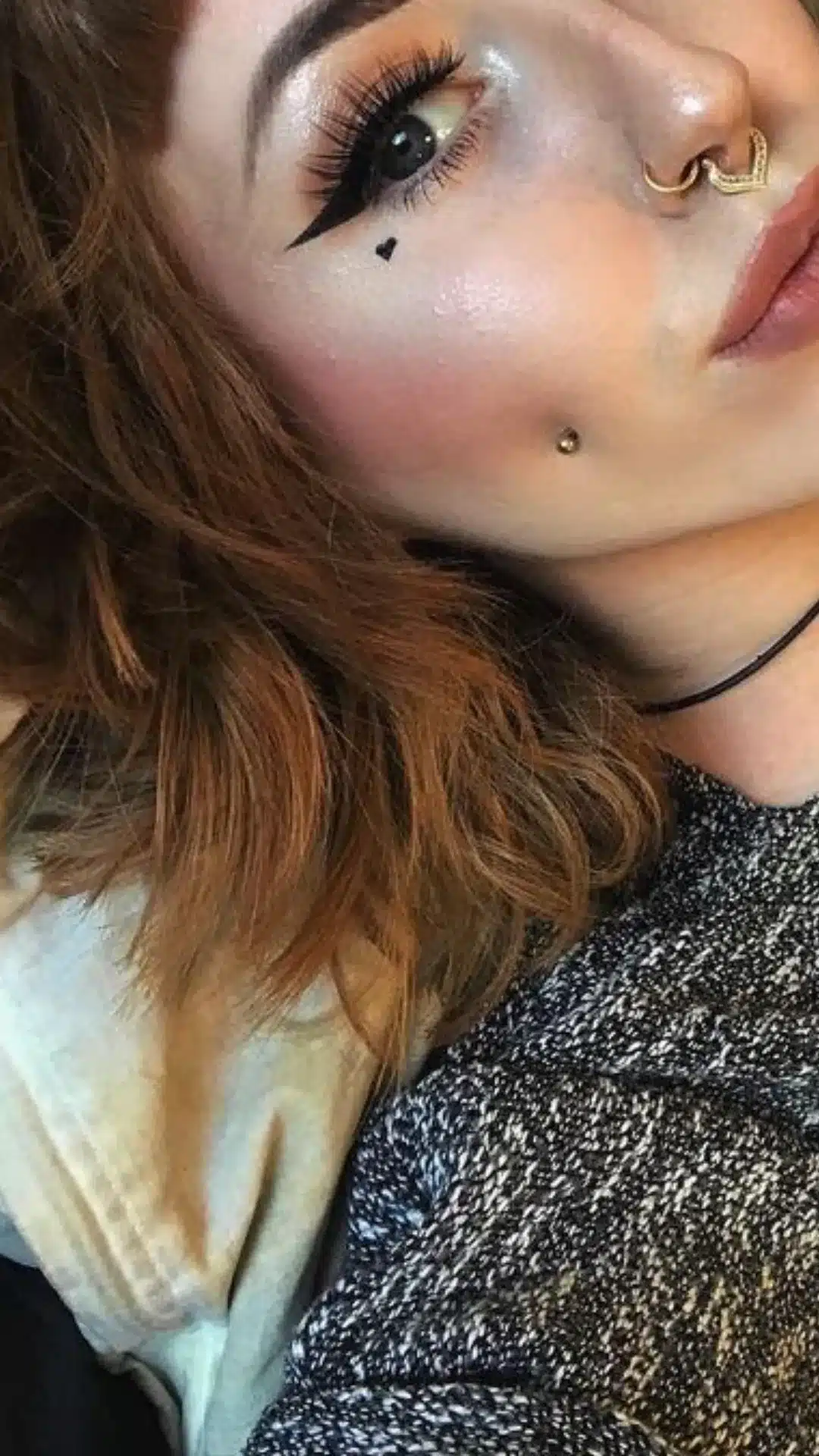 Close-up portrait of a person with winged eyeliner, dramatic eyelashes, a small heart cheek marking, multiple nose piercings, and a cheek stud.