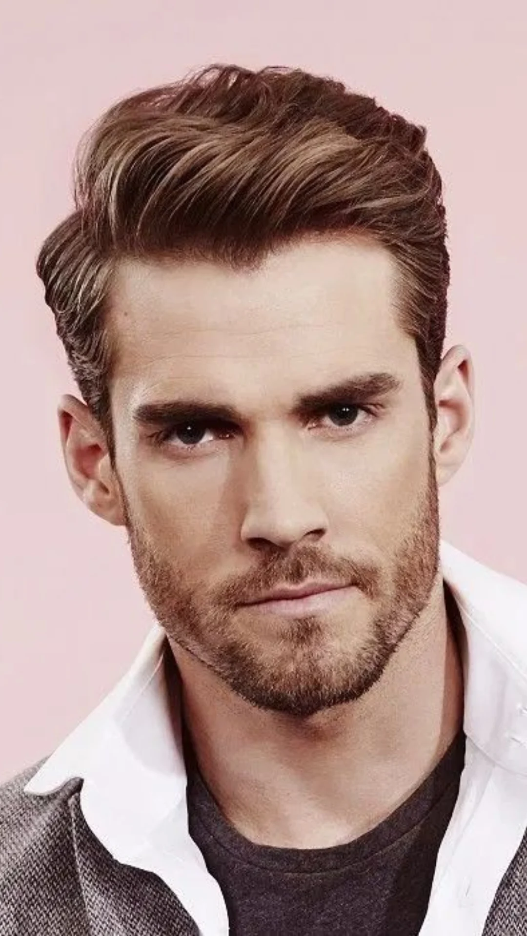 Close-up portrait of a bearded man with styled brown hair wearing a white-collared jacket against a pale pink background.