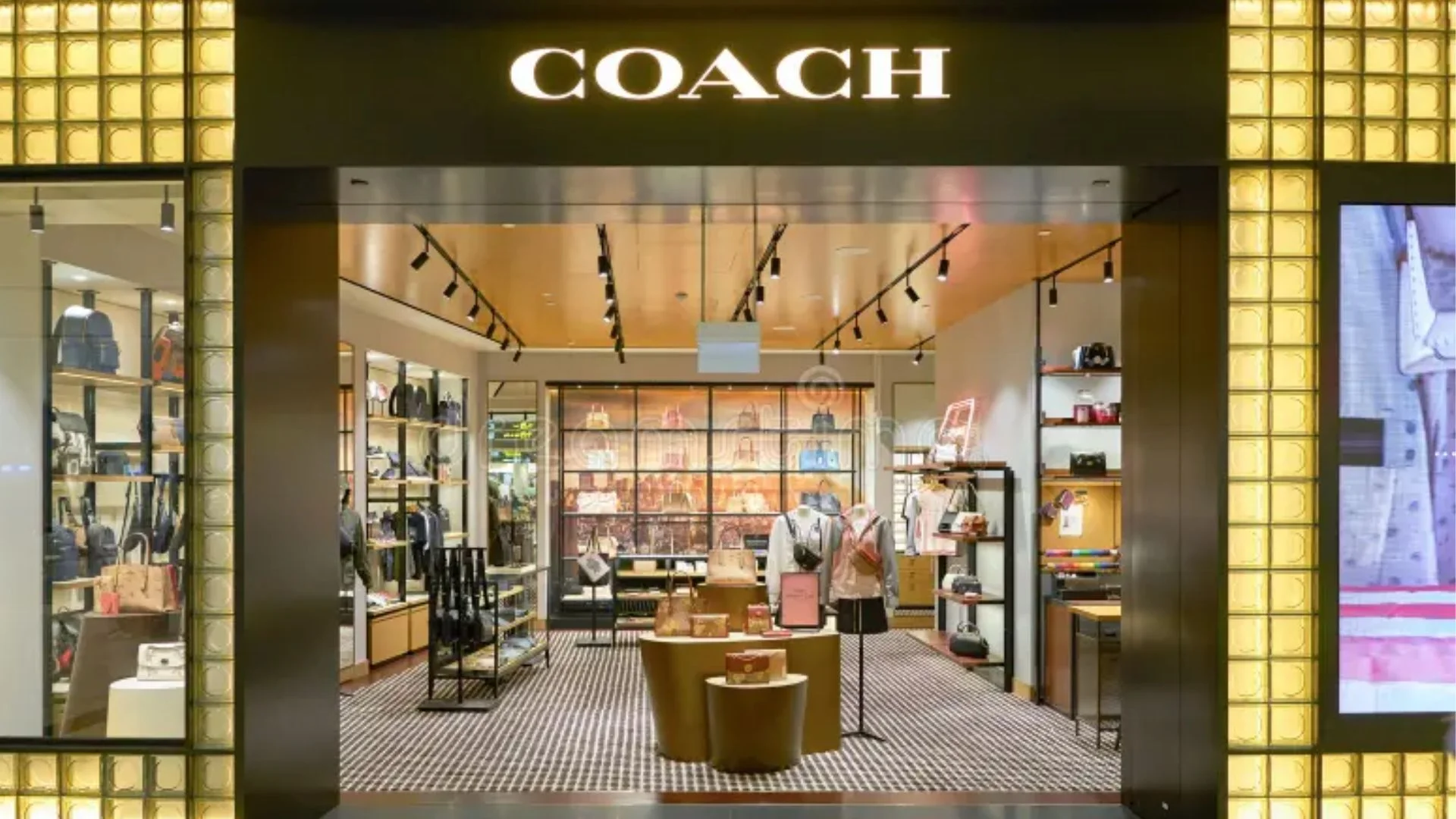 COACH store entrance with illuminated lettering, handbags and clothing displays, mannequins, shelving, and seating inside.