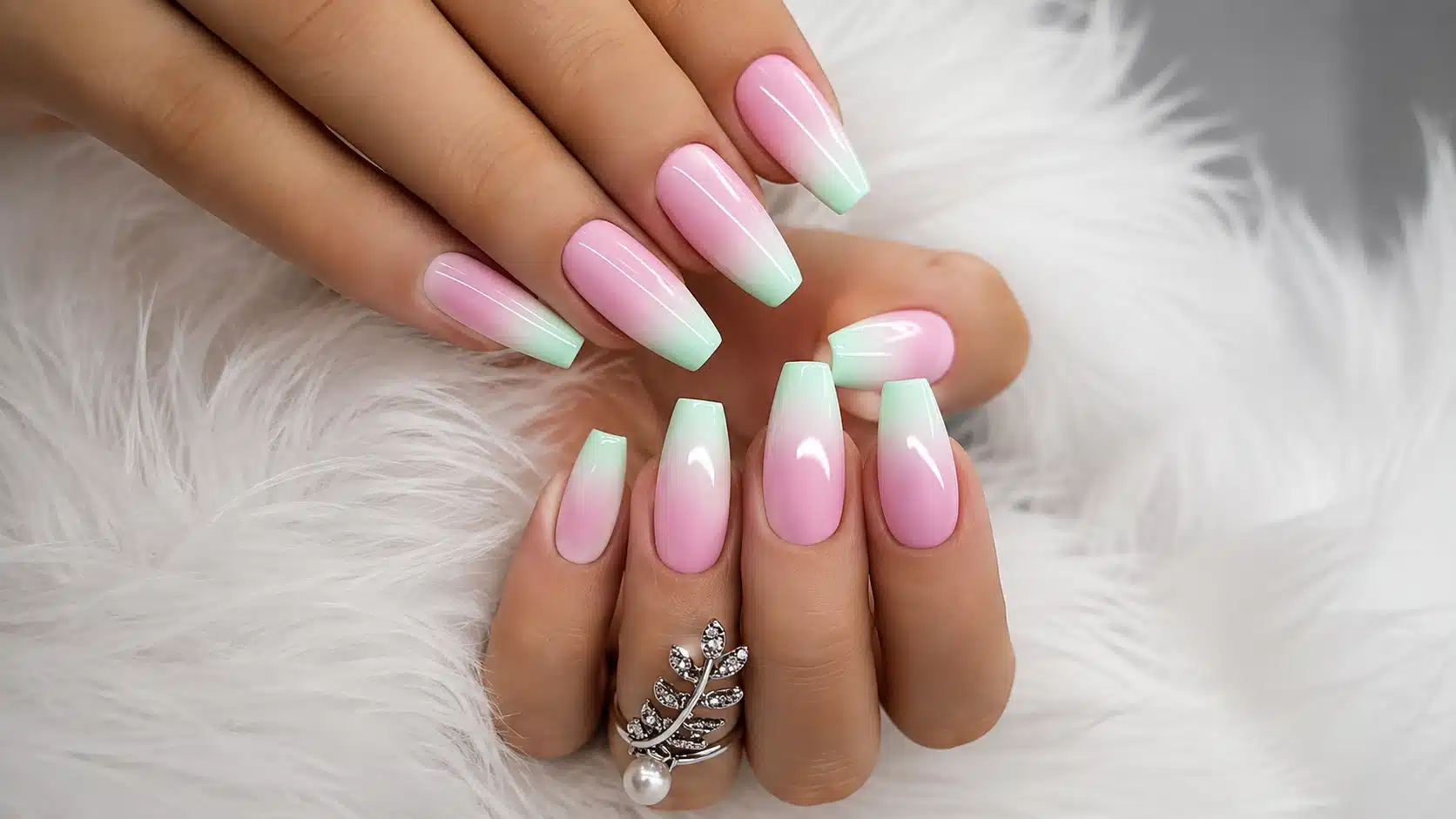 Two hands display glossy pink-to-mint ombre coffin nails with a silver leaf ring against soft white faux fur background