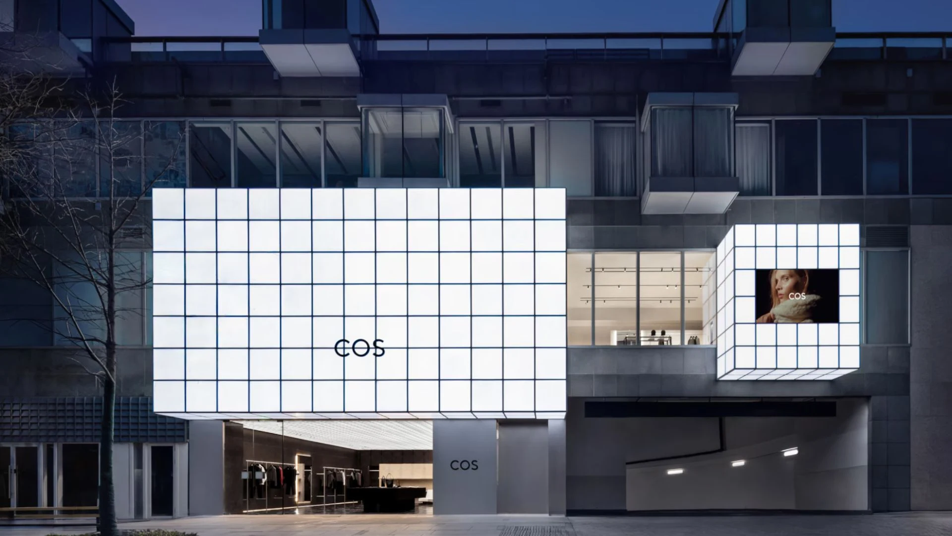 COS storefront with illuminated white grid panels, a fashion display screen, and the modern clothing store entrance beneath a dark apartment building.
