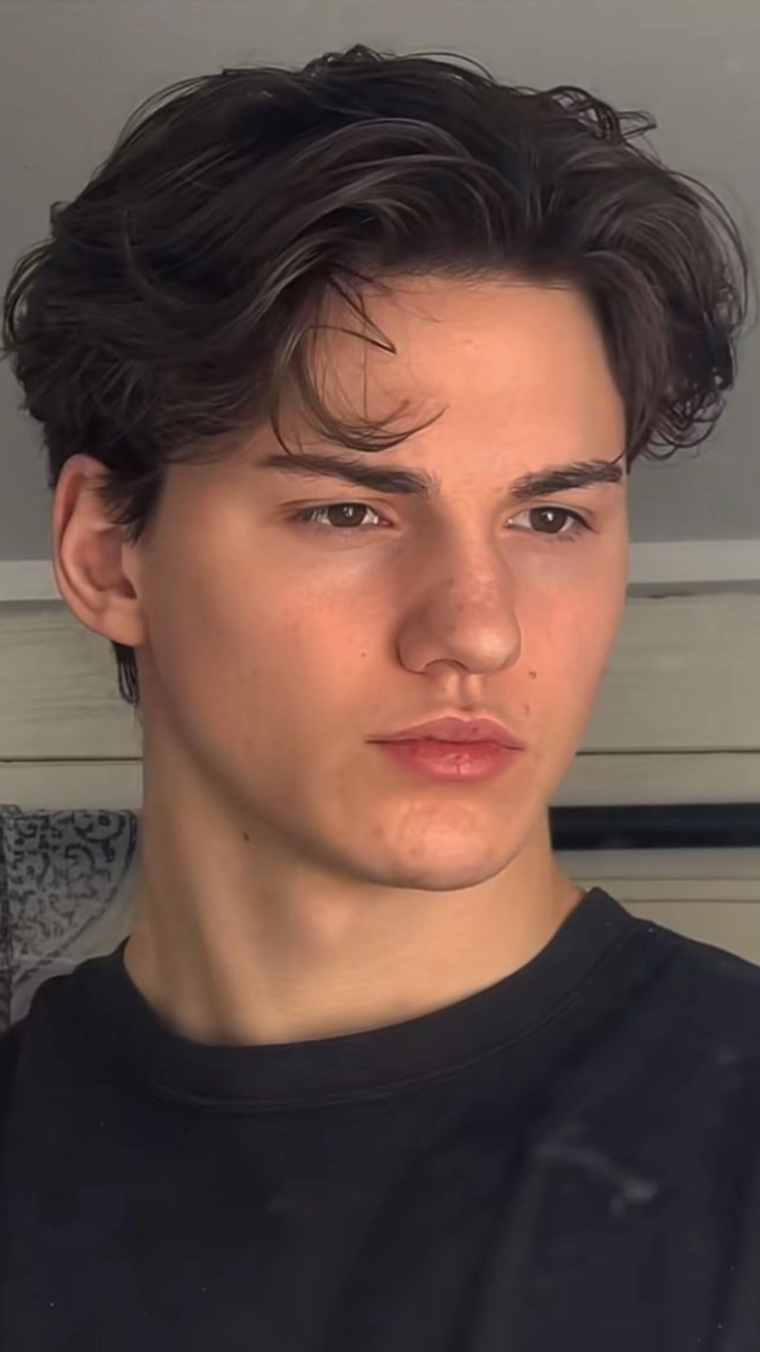 Close-up portrait of a young man with tousled dark hair wearing a black shirt and looking toward the camera indoors.