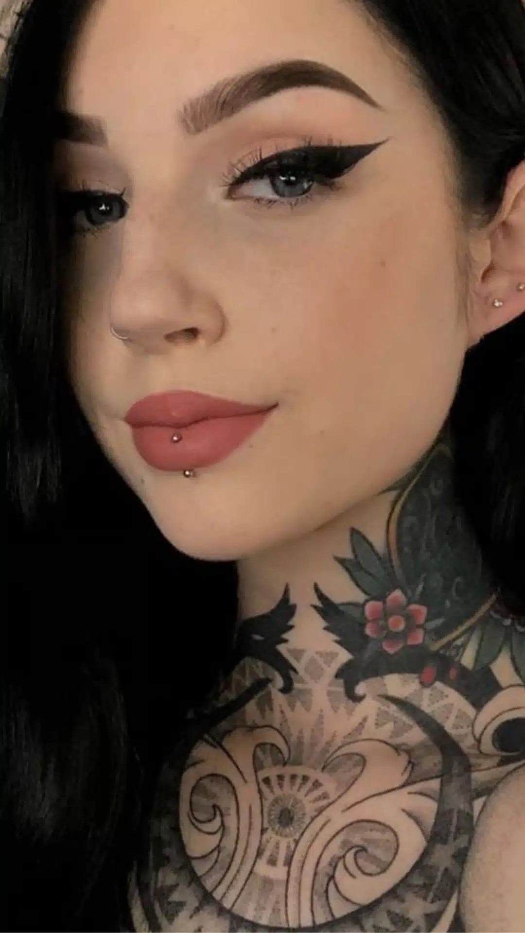Close-up portrait of a woman with black hair, dramatic winged eyeliner, a nose ring, cyber bite lip piercings, and detailed neck tattoos.