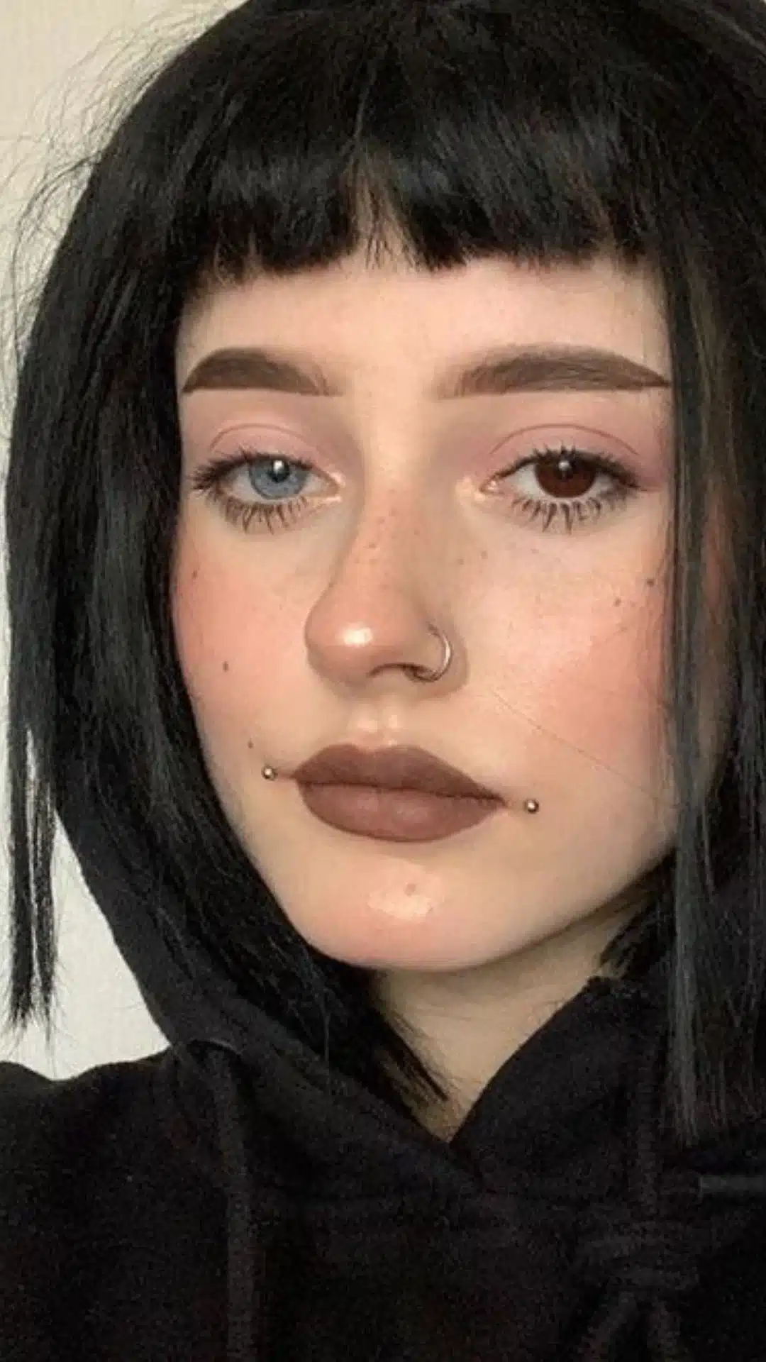 Close-up portrait of a woman with black hair, brown lipstick, a nose ring, and symmetrical Dahlia piercings at the corners of her mouth.